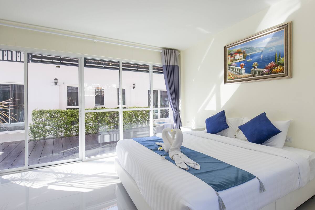A hotel room with a large bed, a painting of a coastal scene, and a balcony with a view of greenery and buildings.