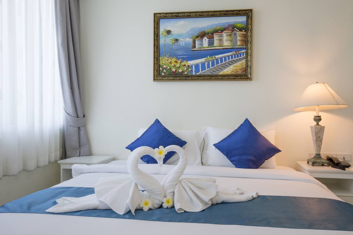A hotel room with a bed, a painting of a coastal scene, a lamp, and a swan-shaped towel arrangement.