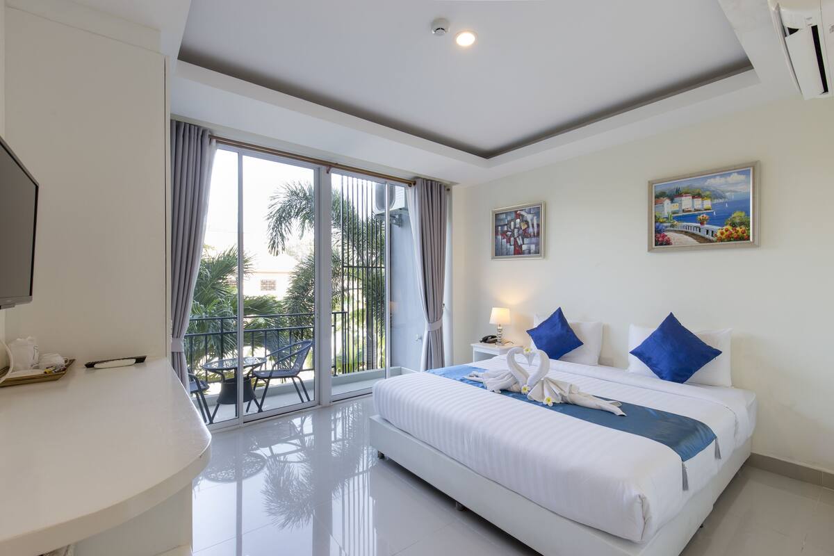 A hotel room with a bed, a desk, a chair, a balcony with a view, and a painting on the wall.