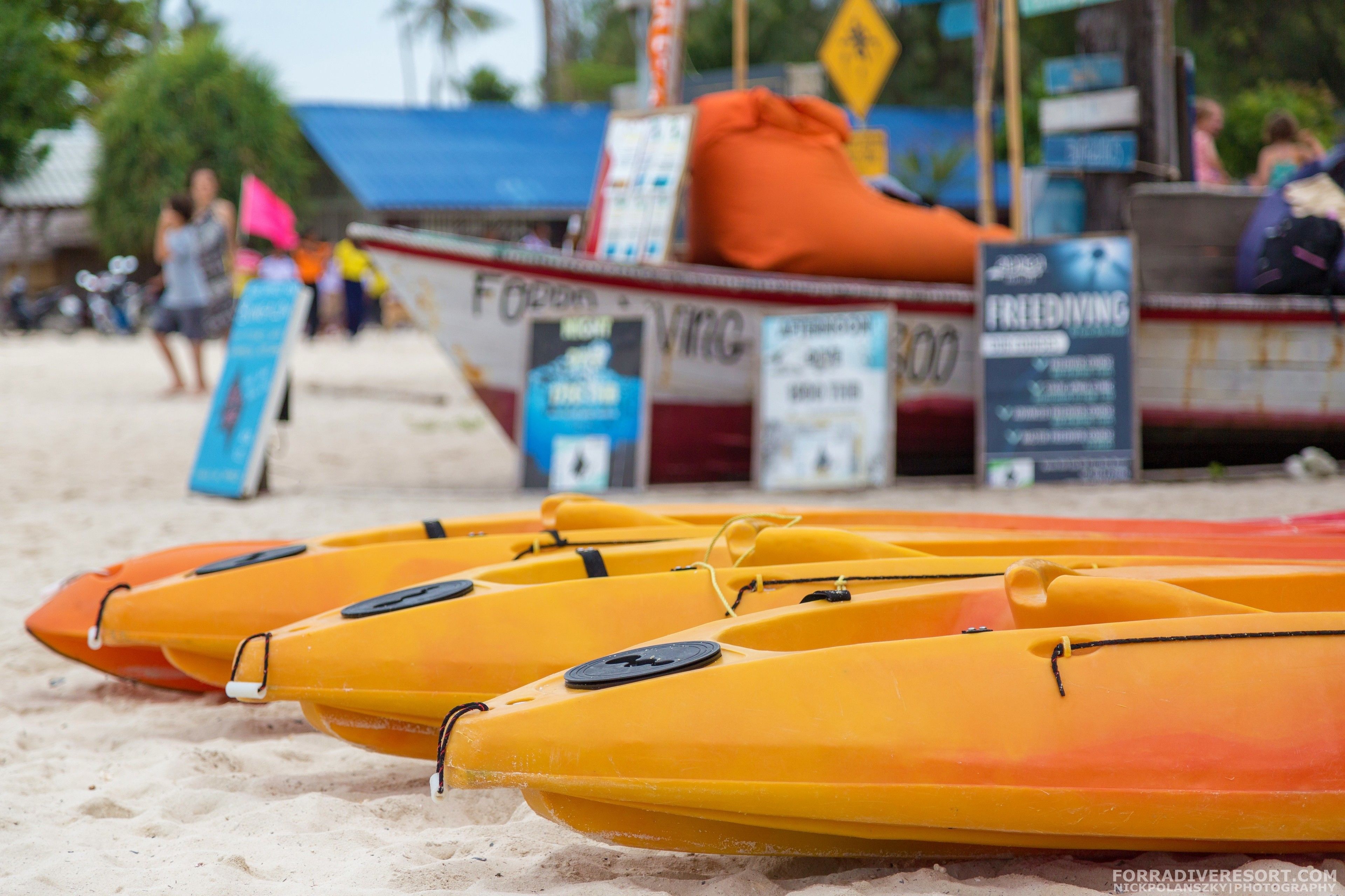 Kayaks lined up on a sandy beach, a boat with a lifebuoy, and a signboard.
