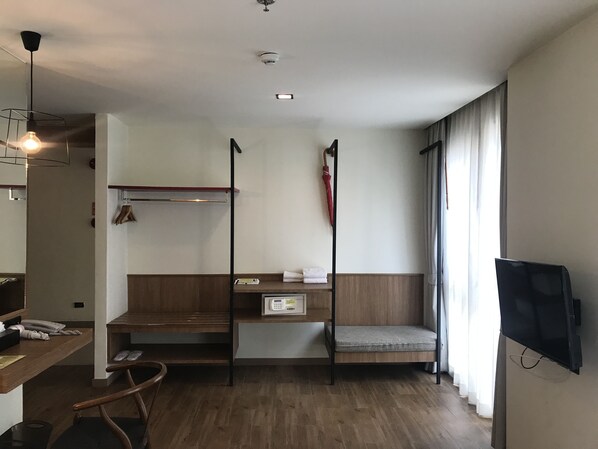A compact hotel room with a wooden desk, a small bed with a grey mattress, a TV mounted on the wall, and a window with curtains.