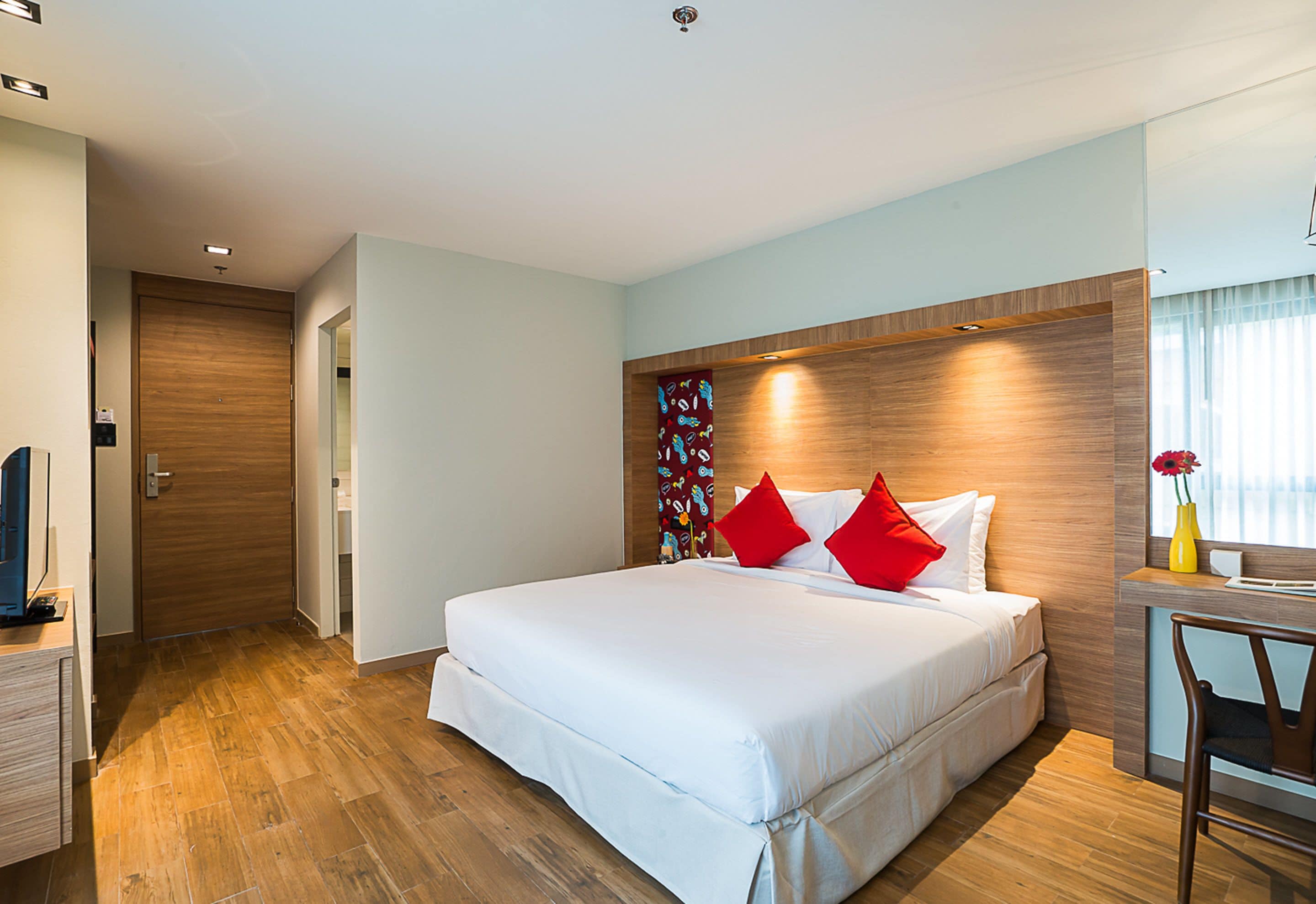 A modern hotel room with a large bed, wooden flooring, a desk with a chair, and a small table with a vase.