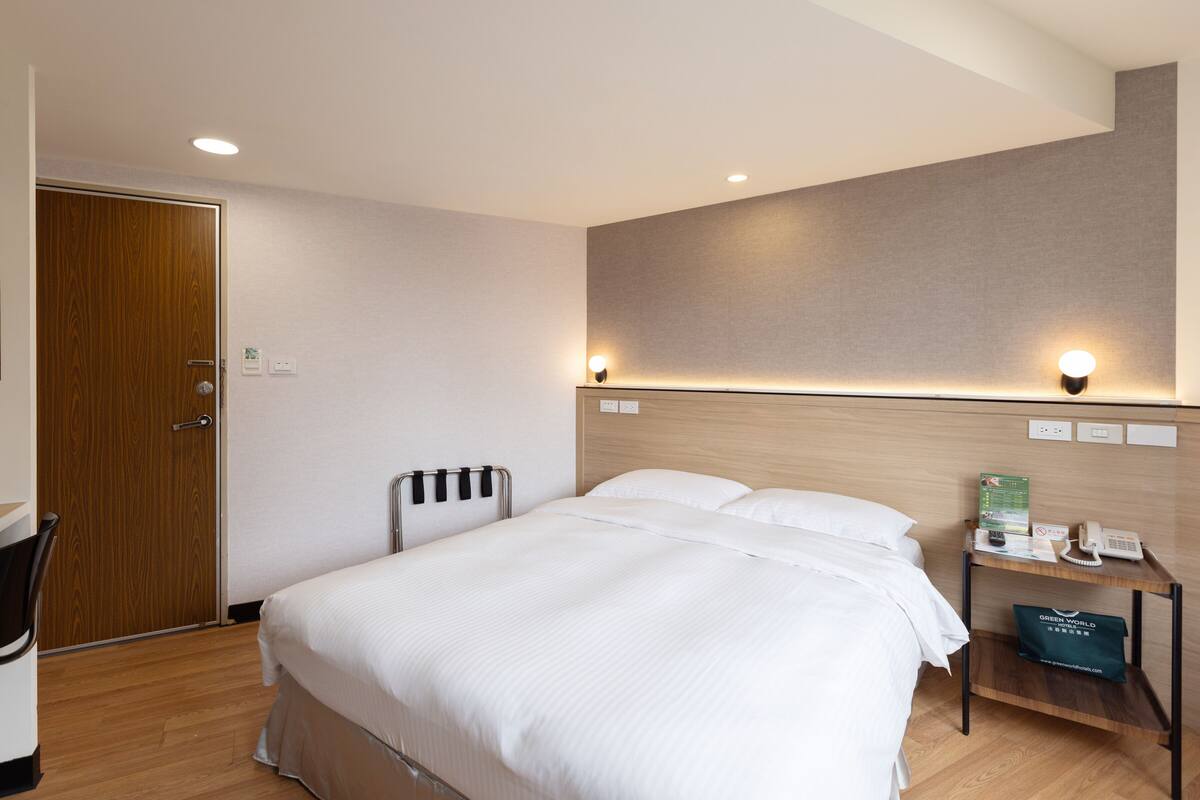 A hotel room with a bed, a desk, a chair, a radiator, and a wall-mounted phone.