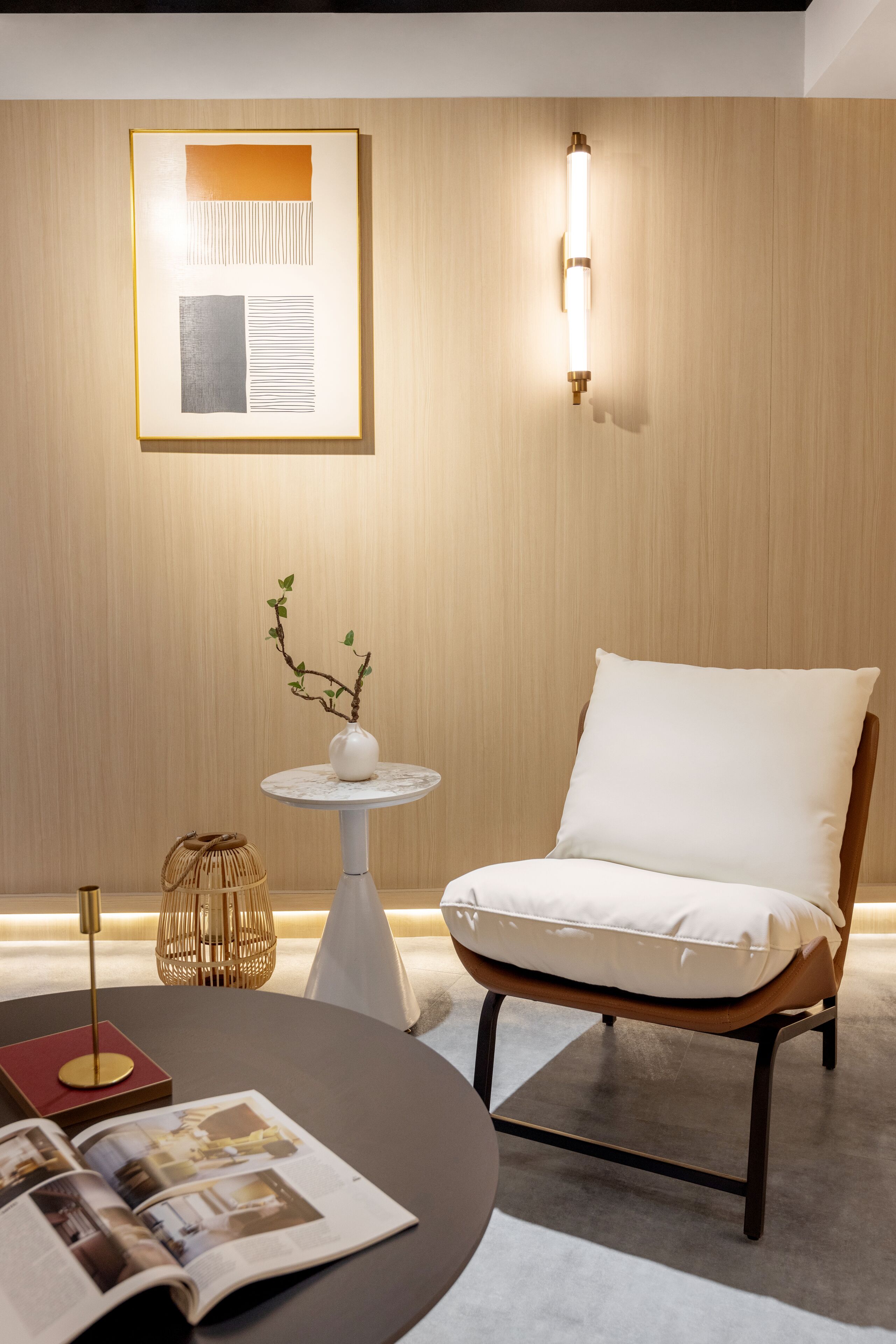 A modern room with a round table, a white armchair, a small round table with a vase, a wall-mounted light, and a framed picture on the wall.