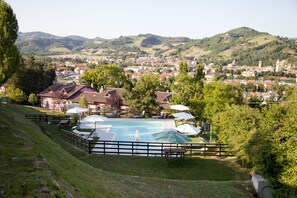 Un espace piscine doté de chaises longues et de parasols, entouré d’une clôture, et offrant une vue sur une ville et des collines en arrière-plan.