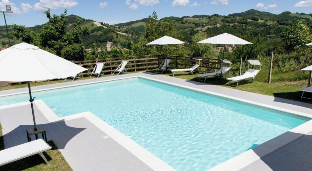 A rectangular swimming pool with clear blue water, surrounded by white lounge chairs and umbrellas, set against a backdrop of green hills and a clear sky.