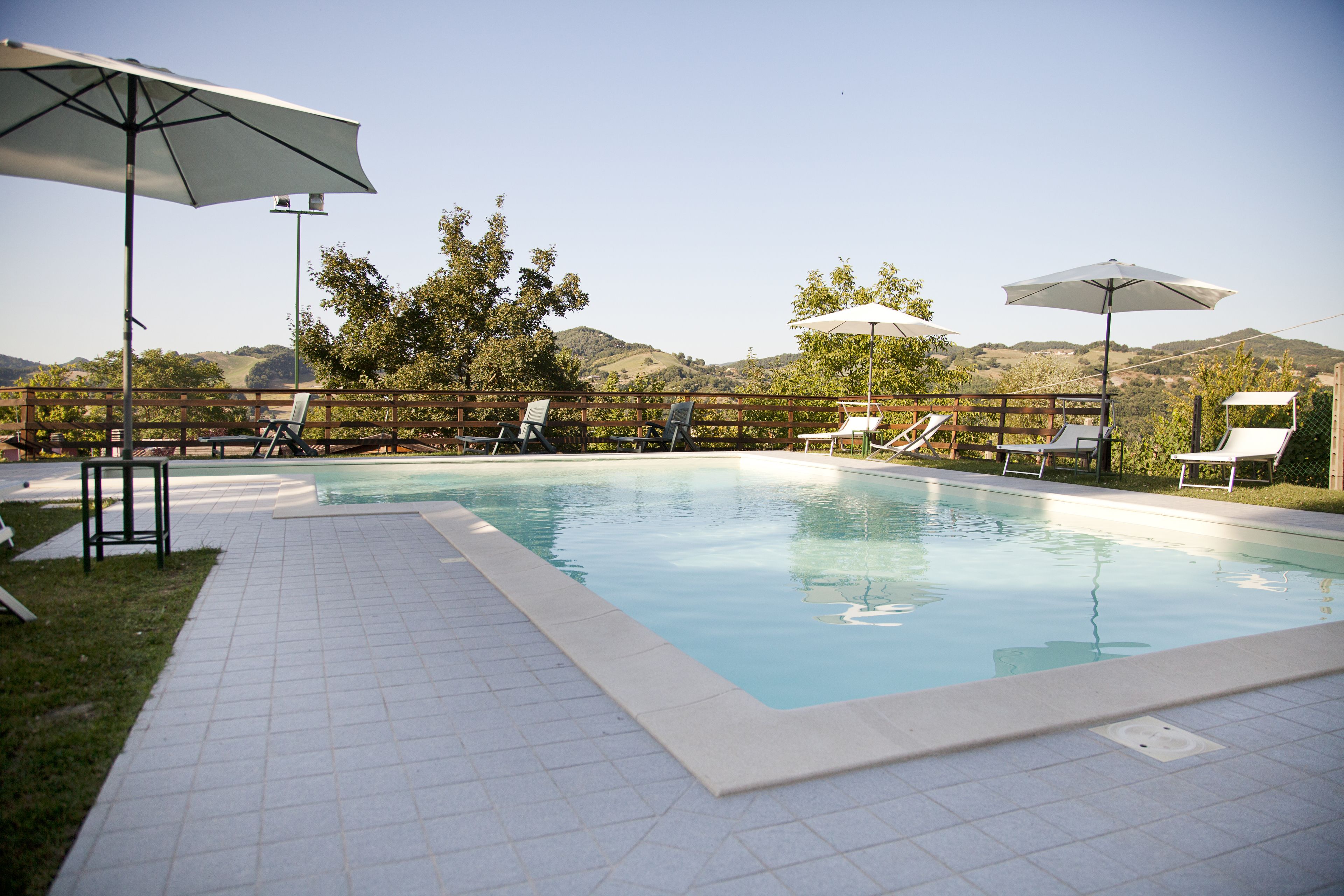 Ein Schwimmbecken mit Fliesenboden, umgeben von Liegestühlen und Sonnenschirmen, vor Hügeln.