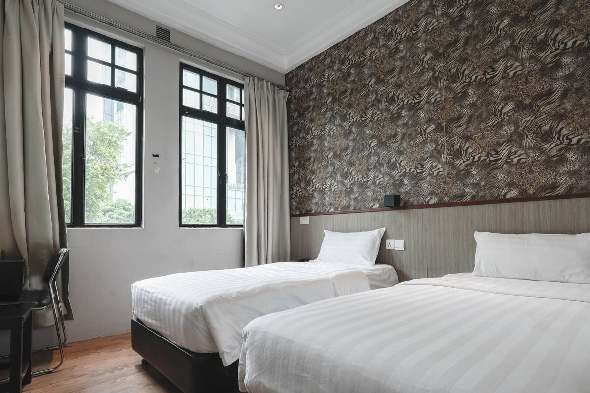 Quarto de hotel com duas camas, uma escrivaninha e uma cadeira. Há duas janelas com cortinas, e uma parede com padrão.