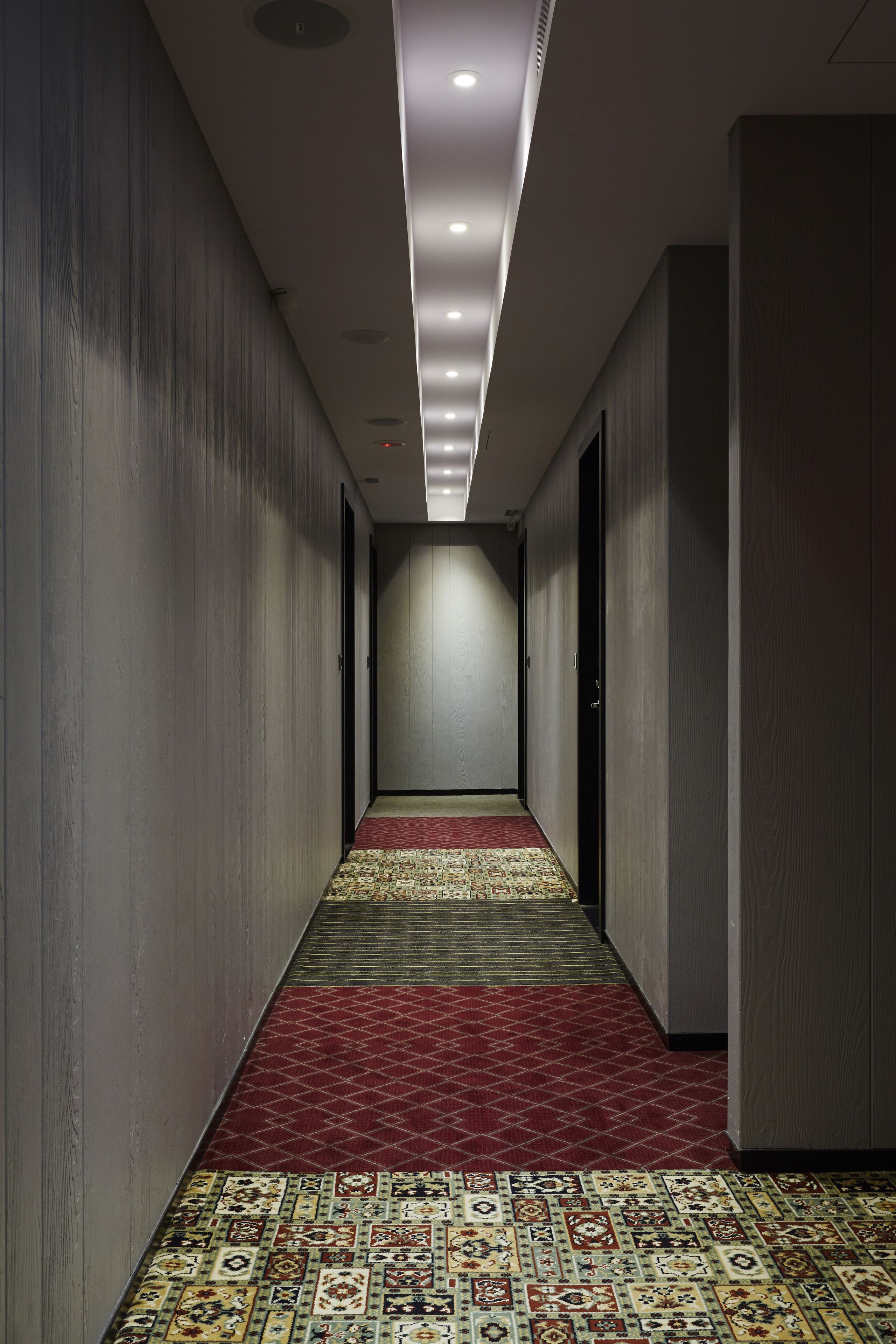 A narrow hallway with patterned carpet, recessed lighting, and plain walls.