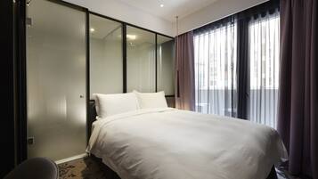 A modern hotel room with a large bed, a sliding glass door, and a view of the city through sheer curtains.