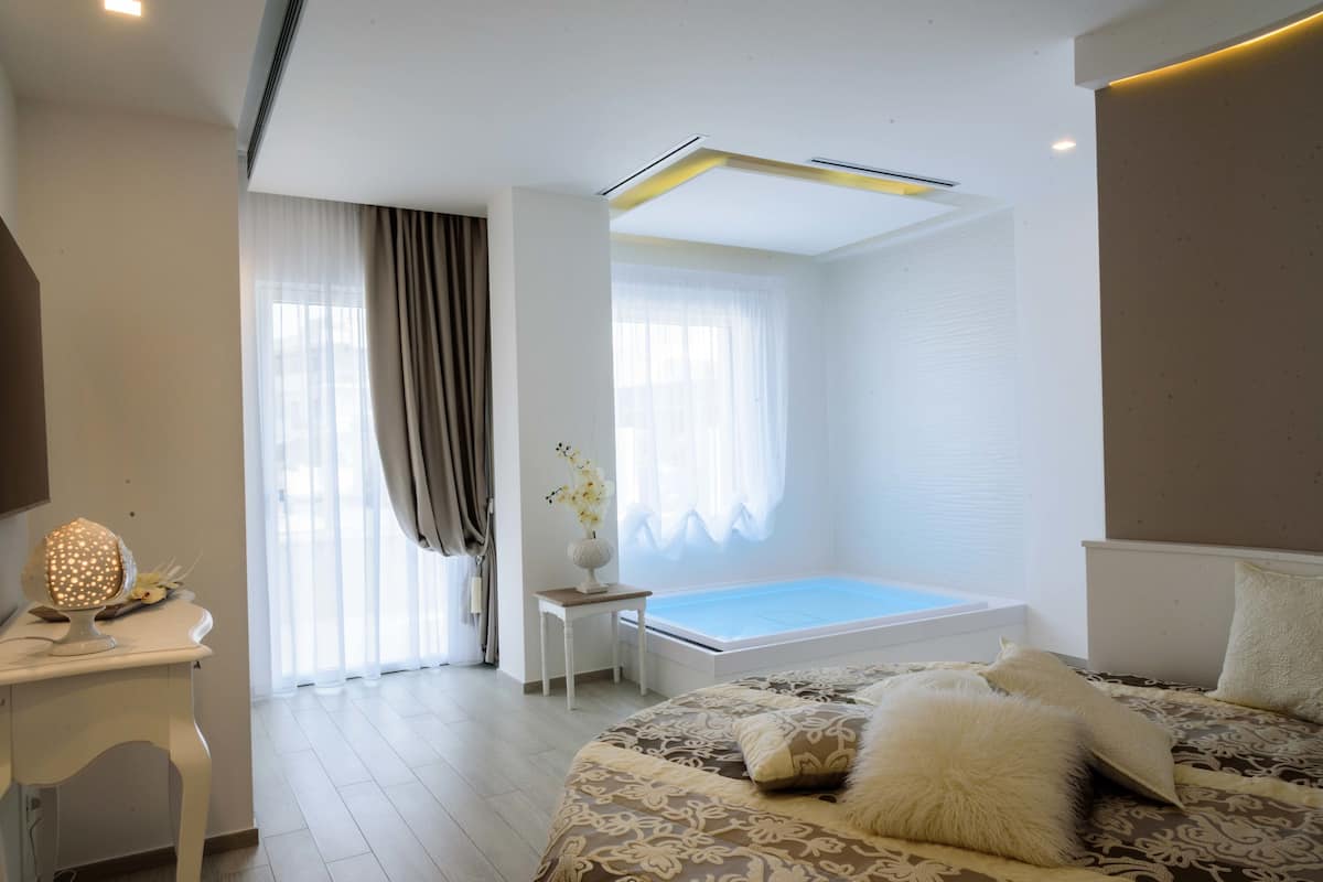 Un dormitorio moderno con una cama grande, una mesita de noche con lámpara, una ventana con cortinas y una bañera empotrada.