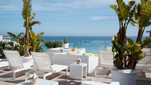 Una terraza en la azotea con mobiliario blanco, una pequeña mesa con un jarrón de flores y plantas grandes en macetas.