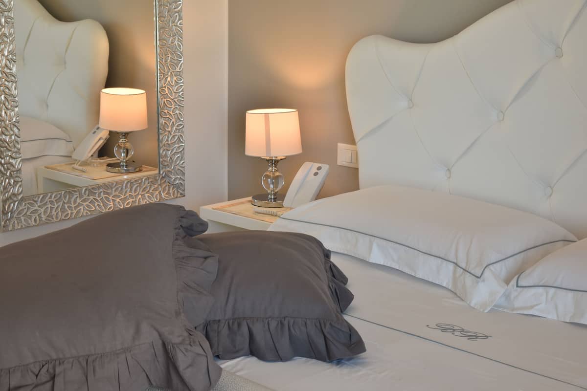 Una cama bien hecha con una manta gris y almohadas, una lámpara de noche, un teléfono y un espejo con un marco decorativo.