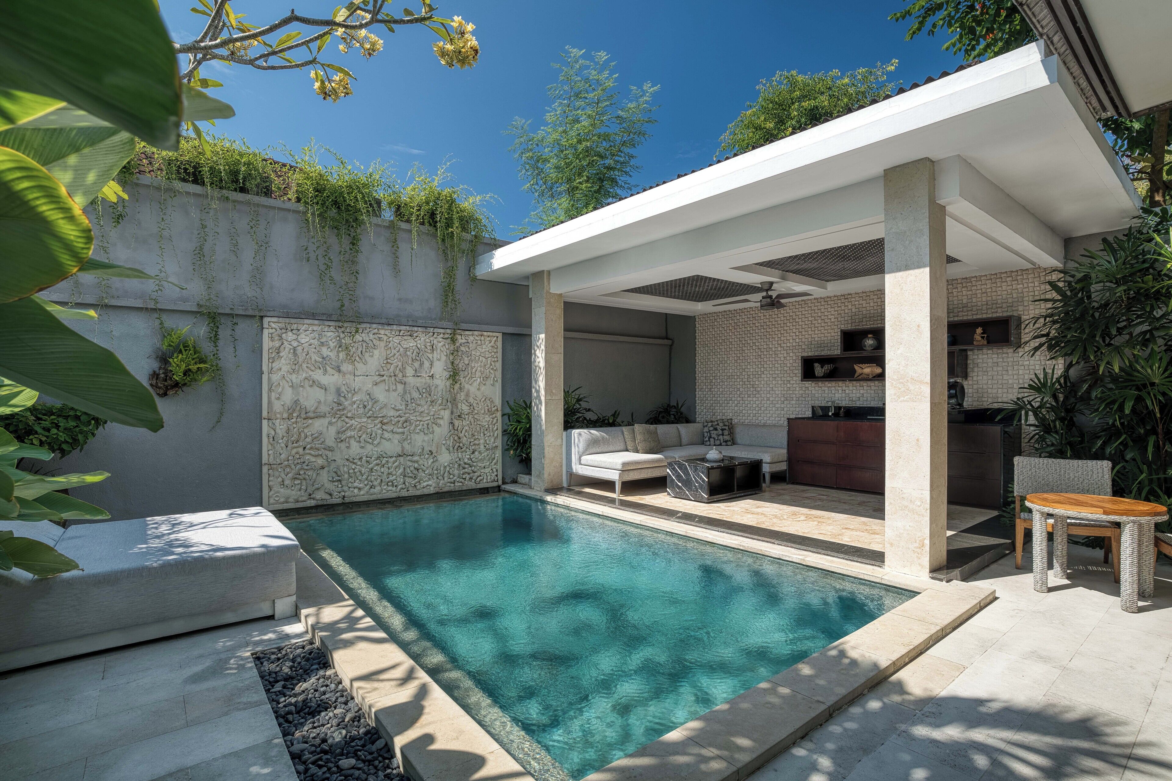 A modern outdoor pool area with a stone wall, a white outdoor seating area, and a covered patio with a kitchen.