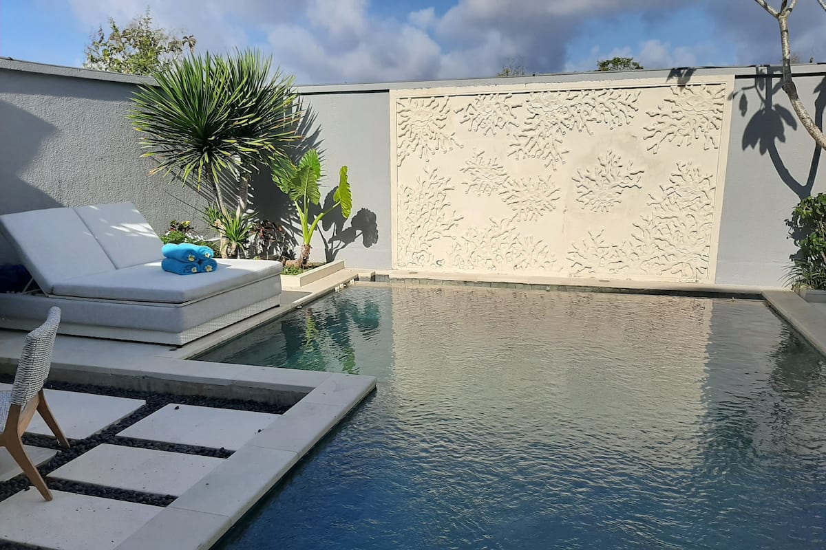 Una piscina con un muro blanco y diseño texturizado, una silla de descanso y una mesita.