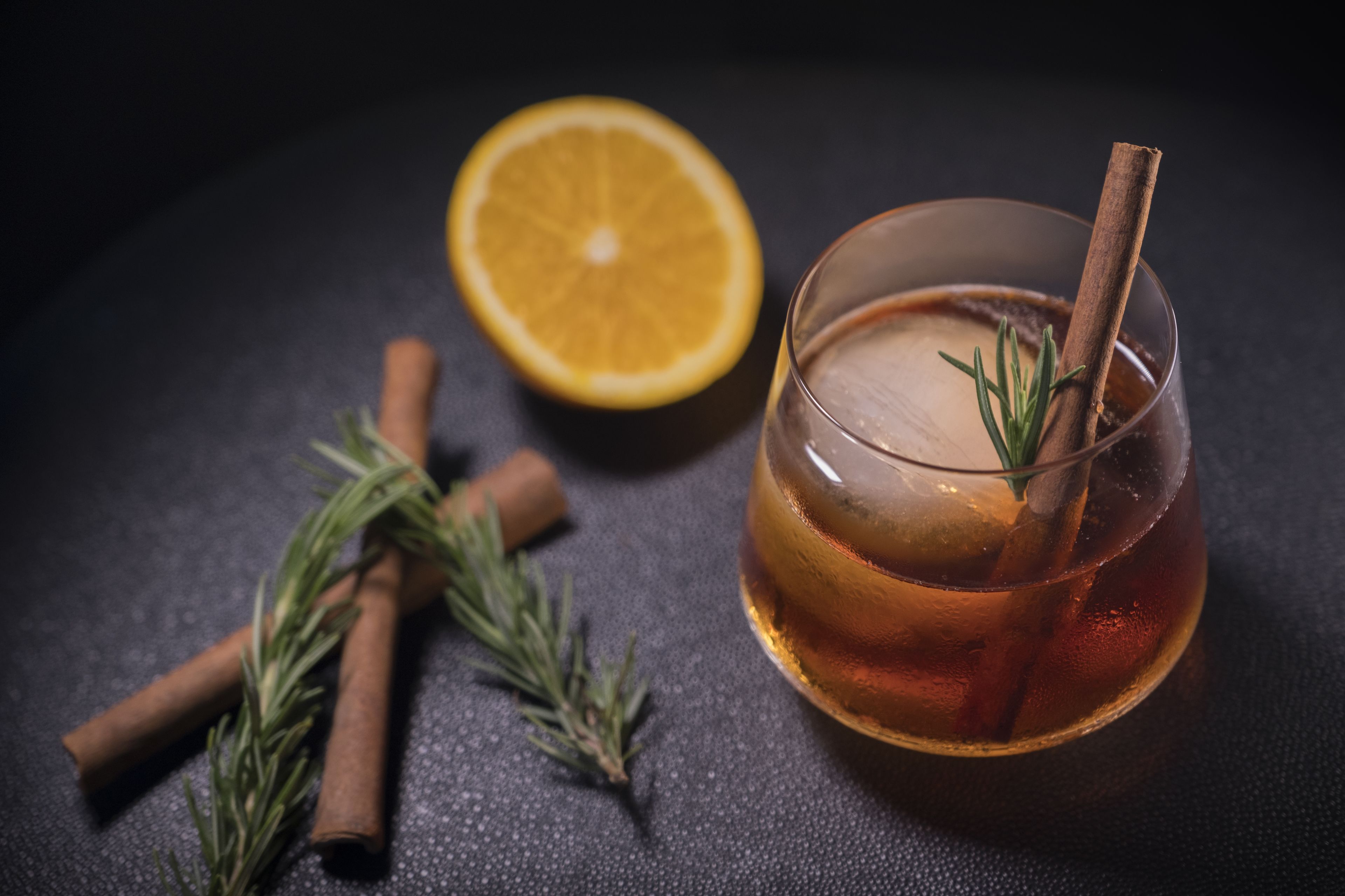 A glass of amber-colored beverage with a cinnamon stick and rosemary garnish, accompanied by a lemon half and cinnamon sticks on a dark surface.