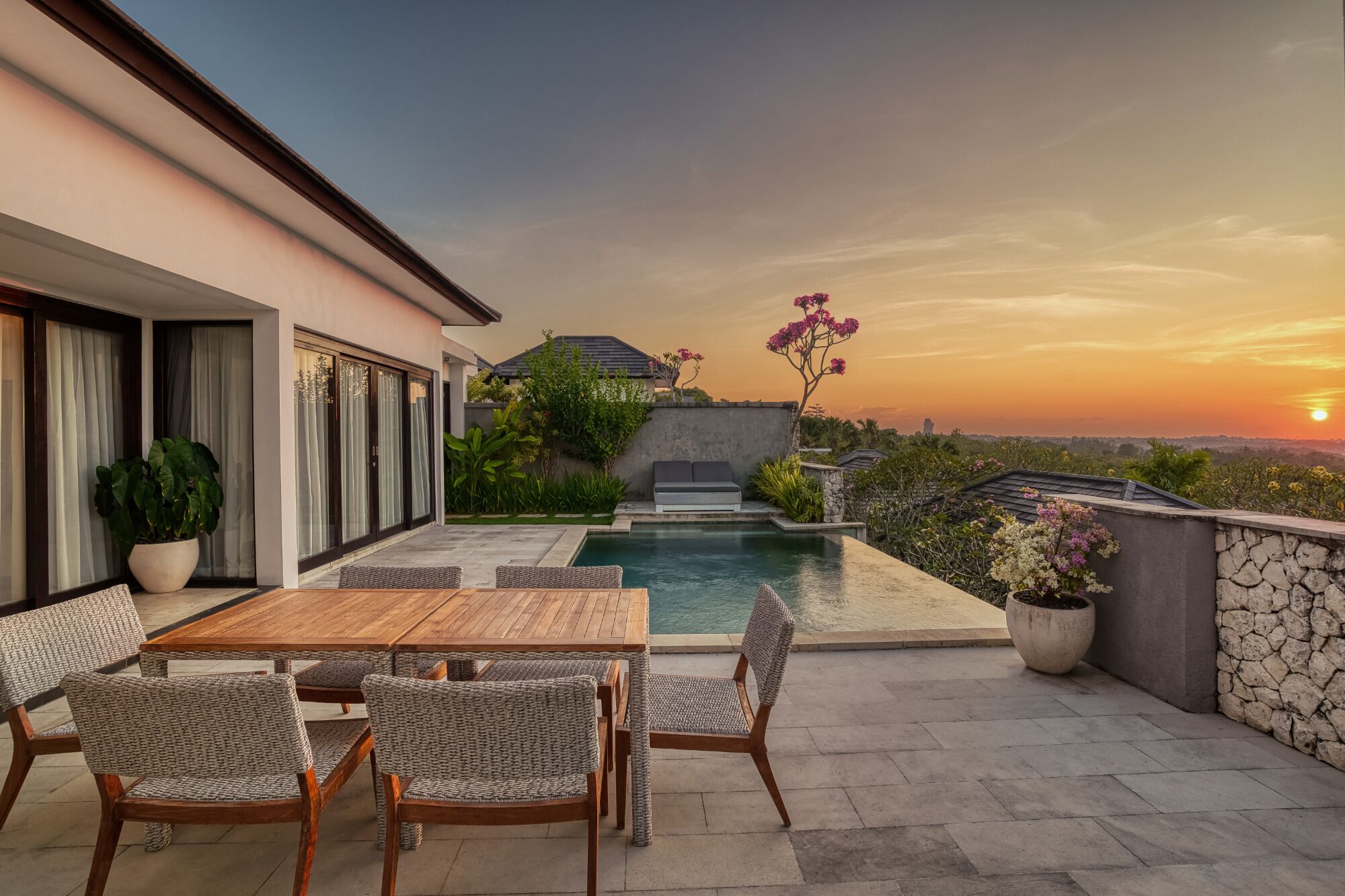 A poolside area with a table and chairs, a stone wall, and a view of the sunset.