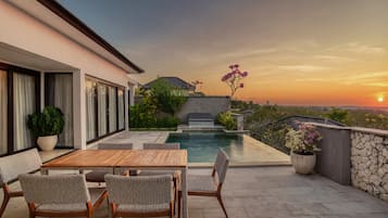 A poolside area with a table and chairs, a stone wall, and a view of the sunset.