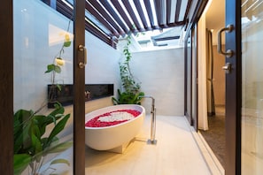 A modern bathroom with a freestanding bathtub filled with rose petals, a glass shower enclosure, and a potted plant.
