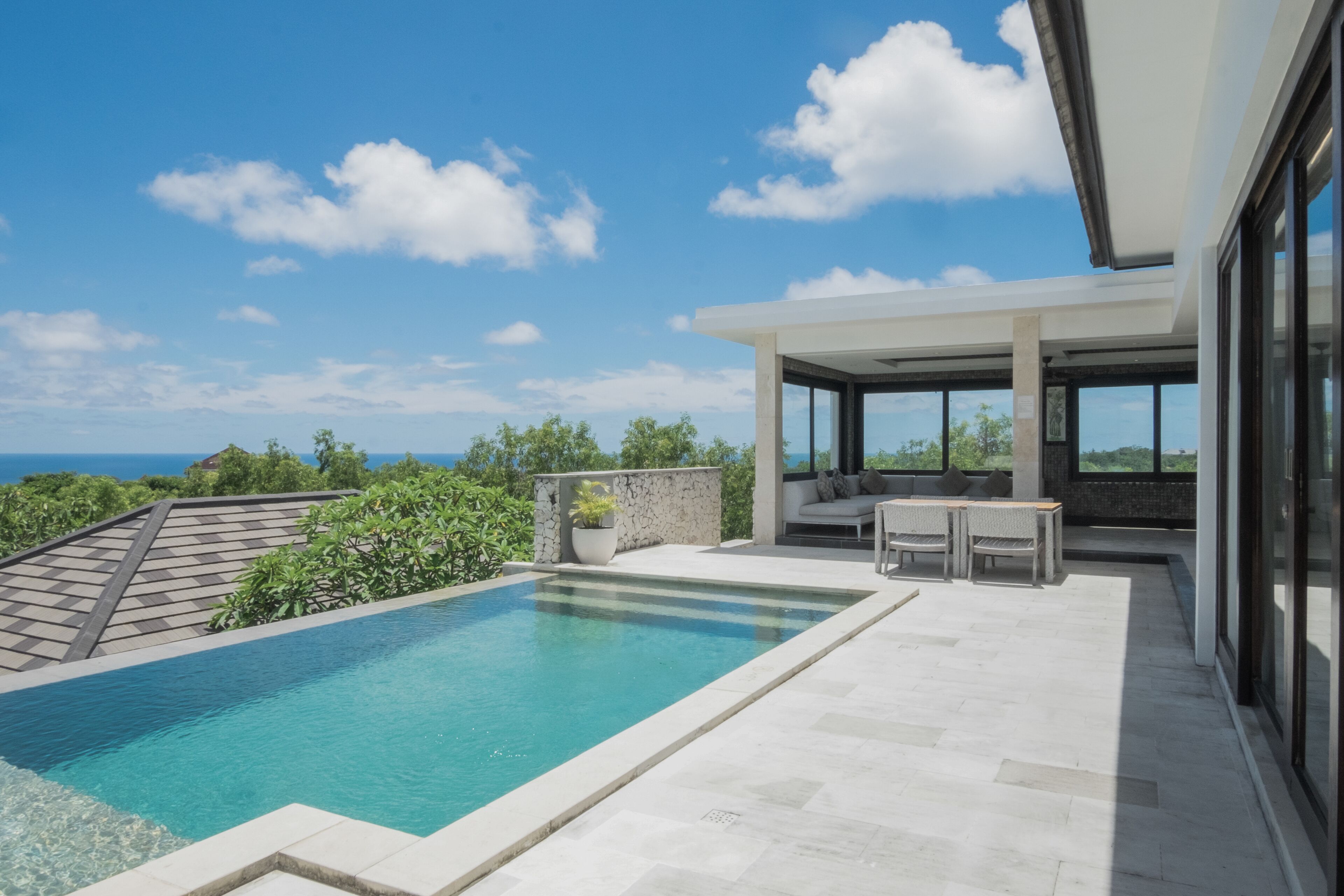 A modern outdoor pool area with a clear view of the ocean, a stone wall, and a seating area with a table and chairs.