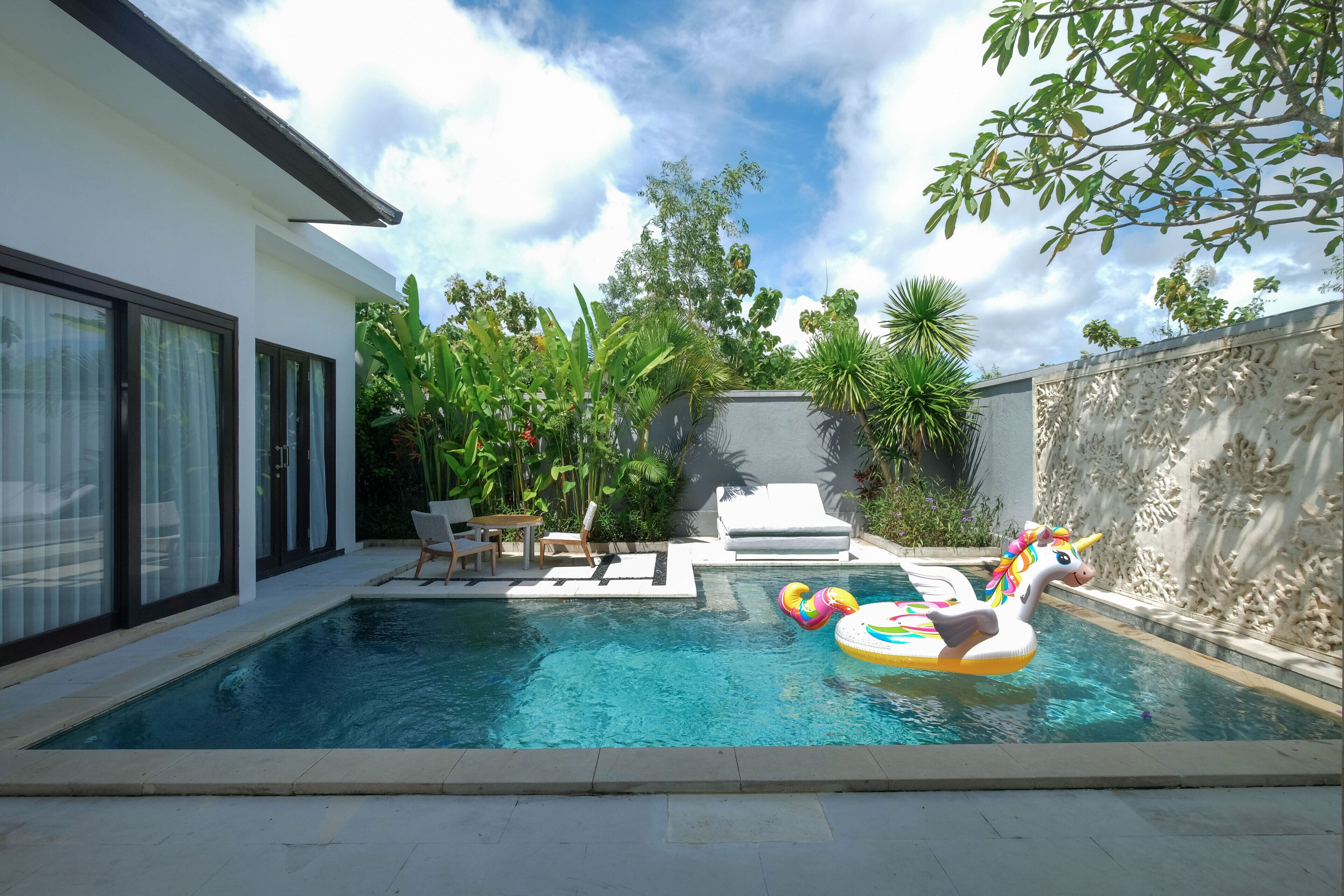 Una piscina con un flotador con forma de unicornio, rodeada de exuberante vegetación y una casa moderna.