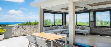 A modern outdoor patio with a dining table and chairs, a sofa, and large windows offering a view of the ocean.