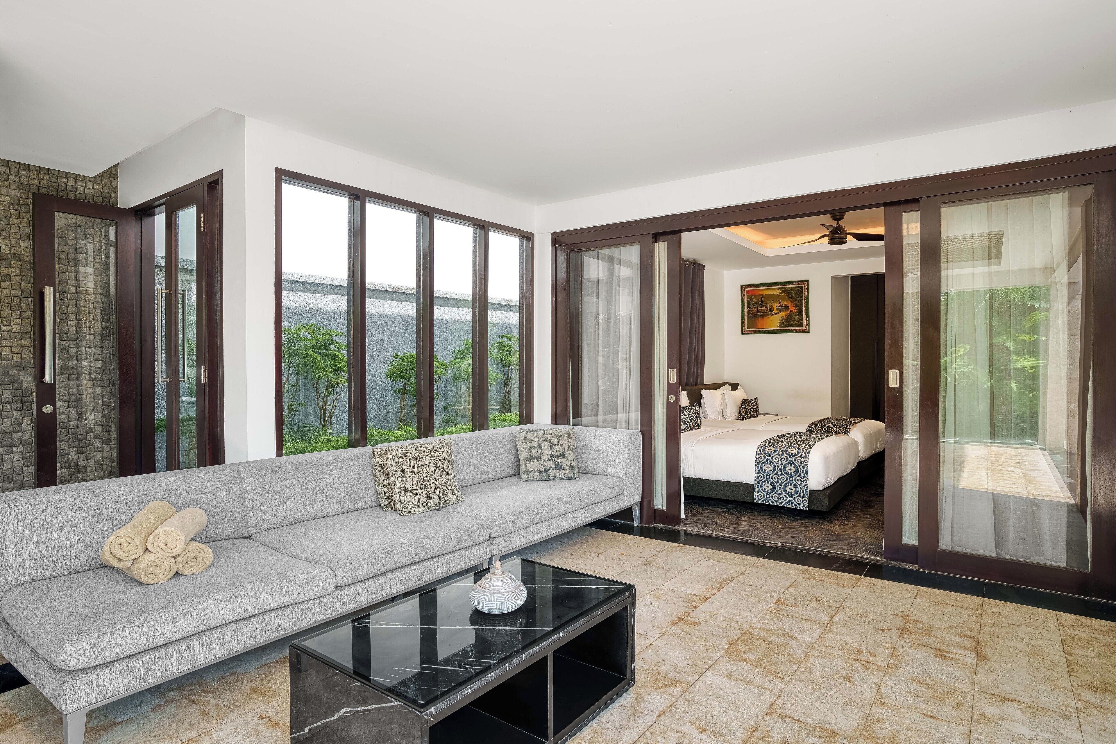 A modern hotel room with a grey sofa, a bed with patterned bedding, and a large window offering a view of greenery.