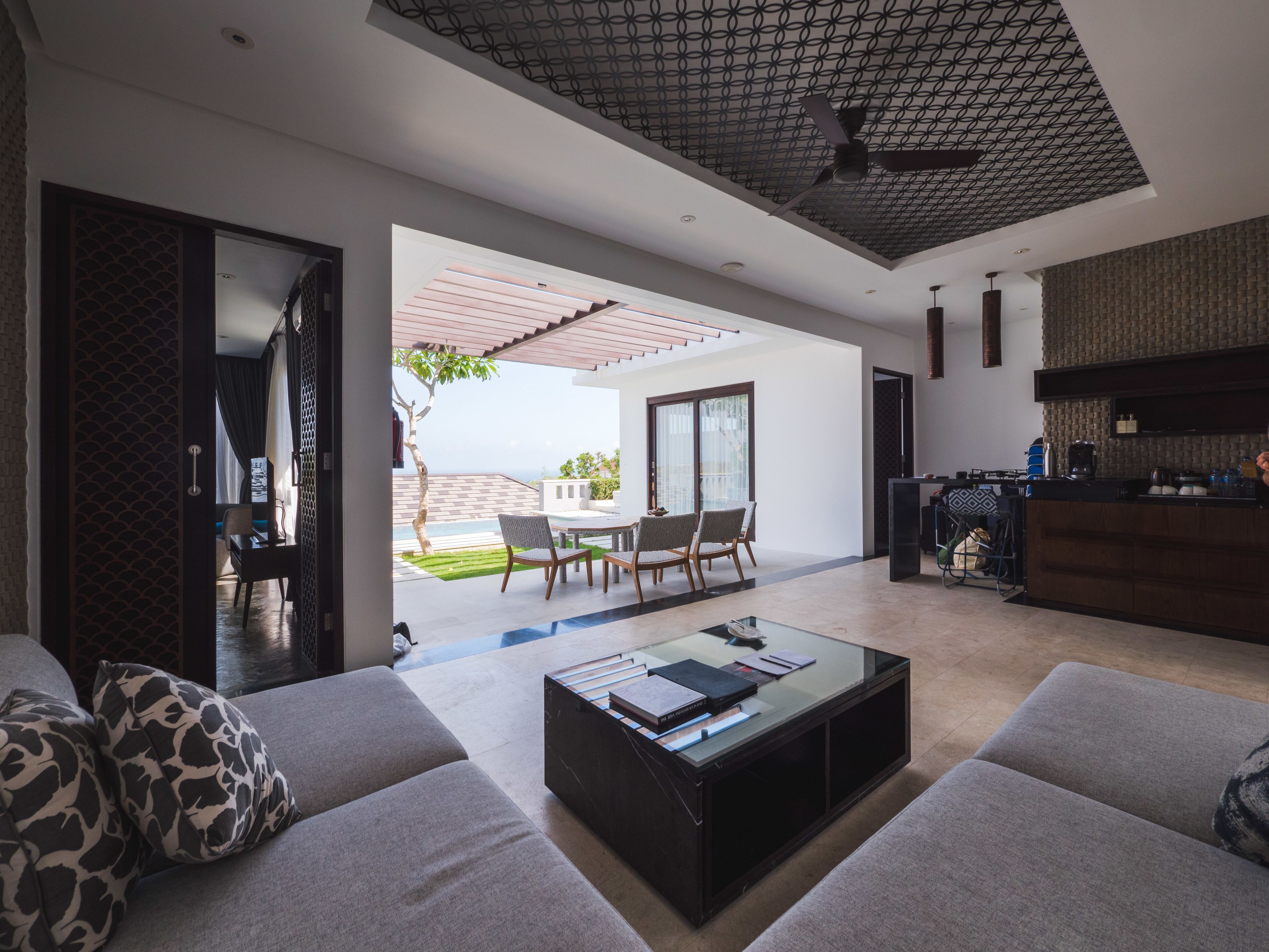 A modern living room with a sofa, coffee table, and dining area. There is a kitchen area with a counter and appliances. The room has a ceiling fan and a view of the outdoors.
