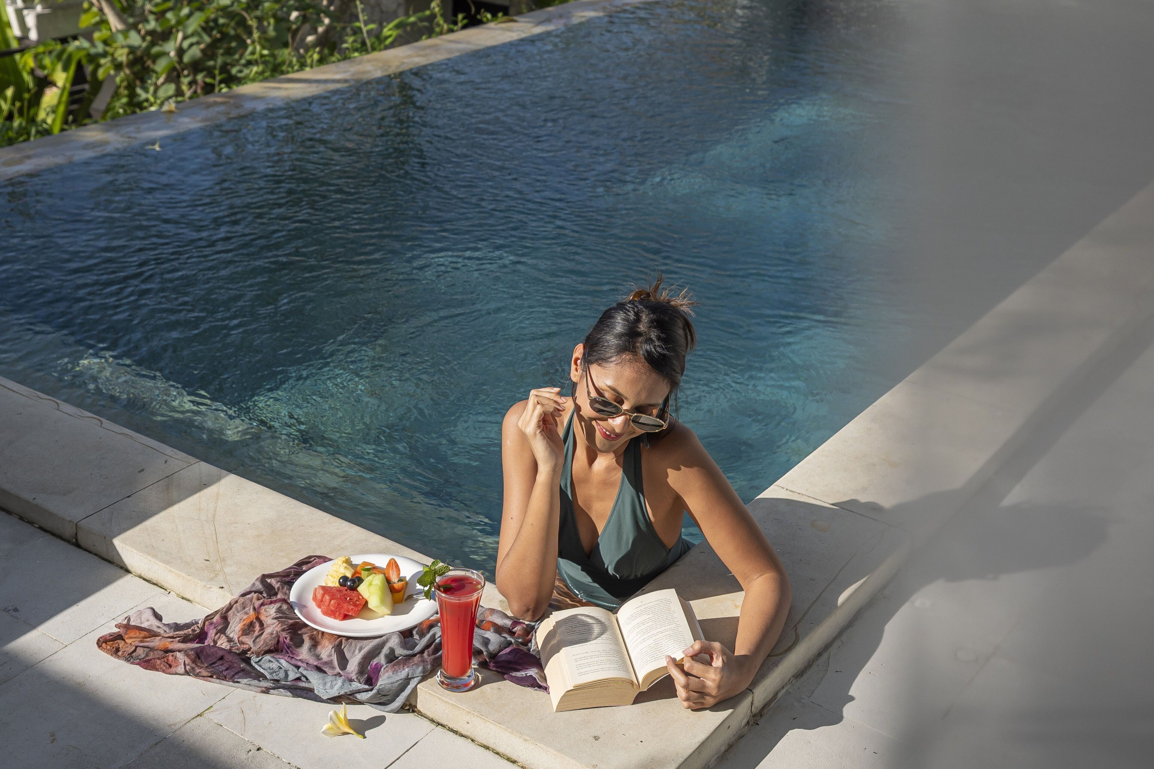 Une personne est assise au bord d’une piscine, un livre à la main, et a devant elle une assiette de nourriture et une boisson.