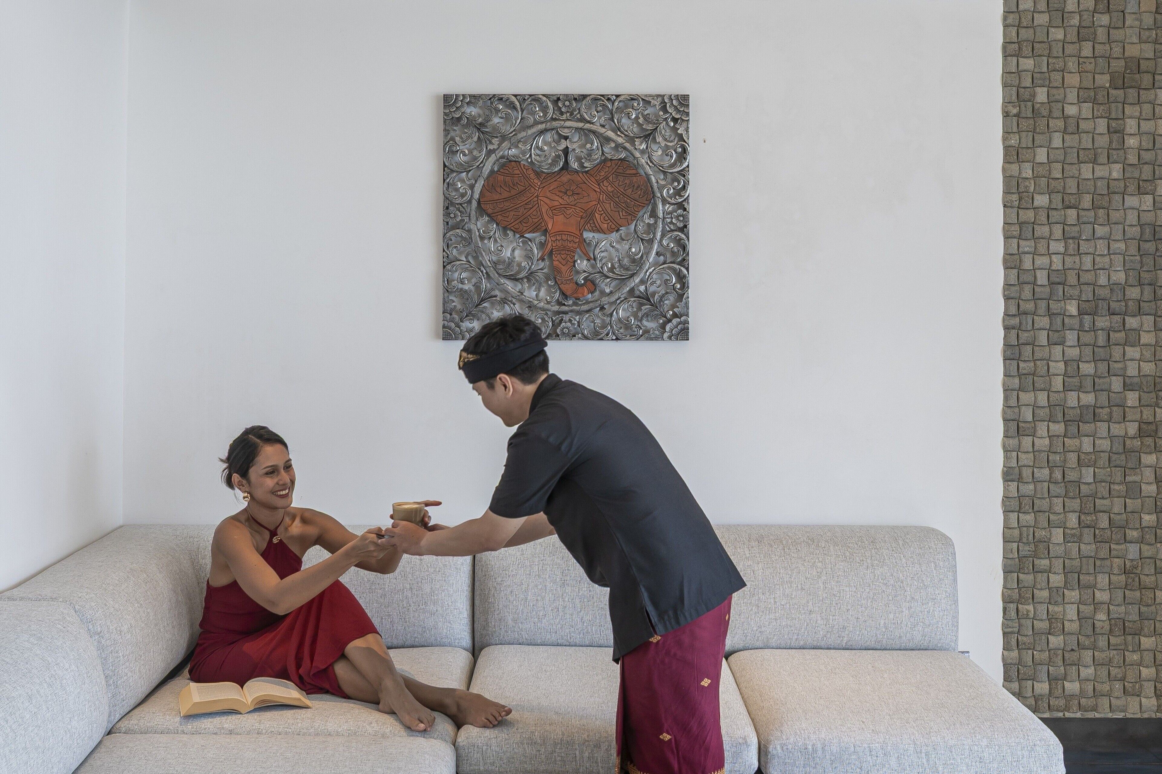 A person sitting on a grey sofa with a book, another person standing beside them, and a wall with a decorative piece.