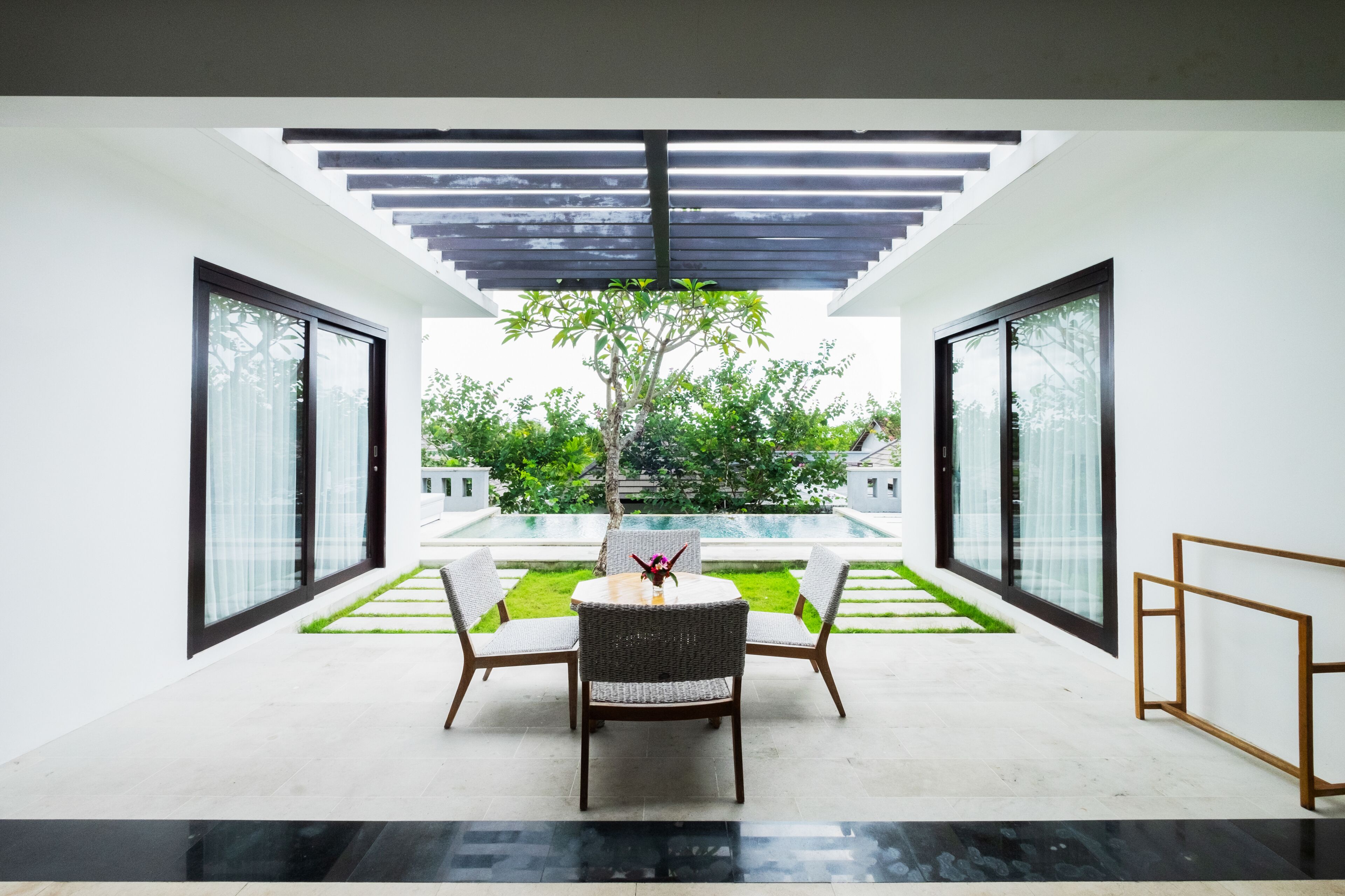 A modern outdoor seating area with a table and chairs, a pergola overhead, and a view of a garden and building in the background.