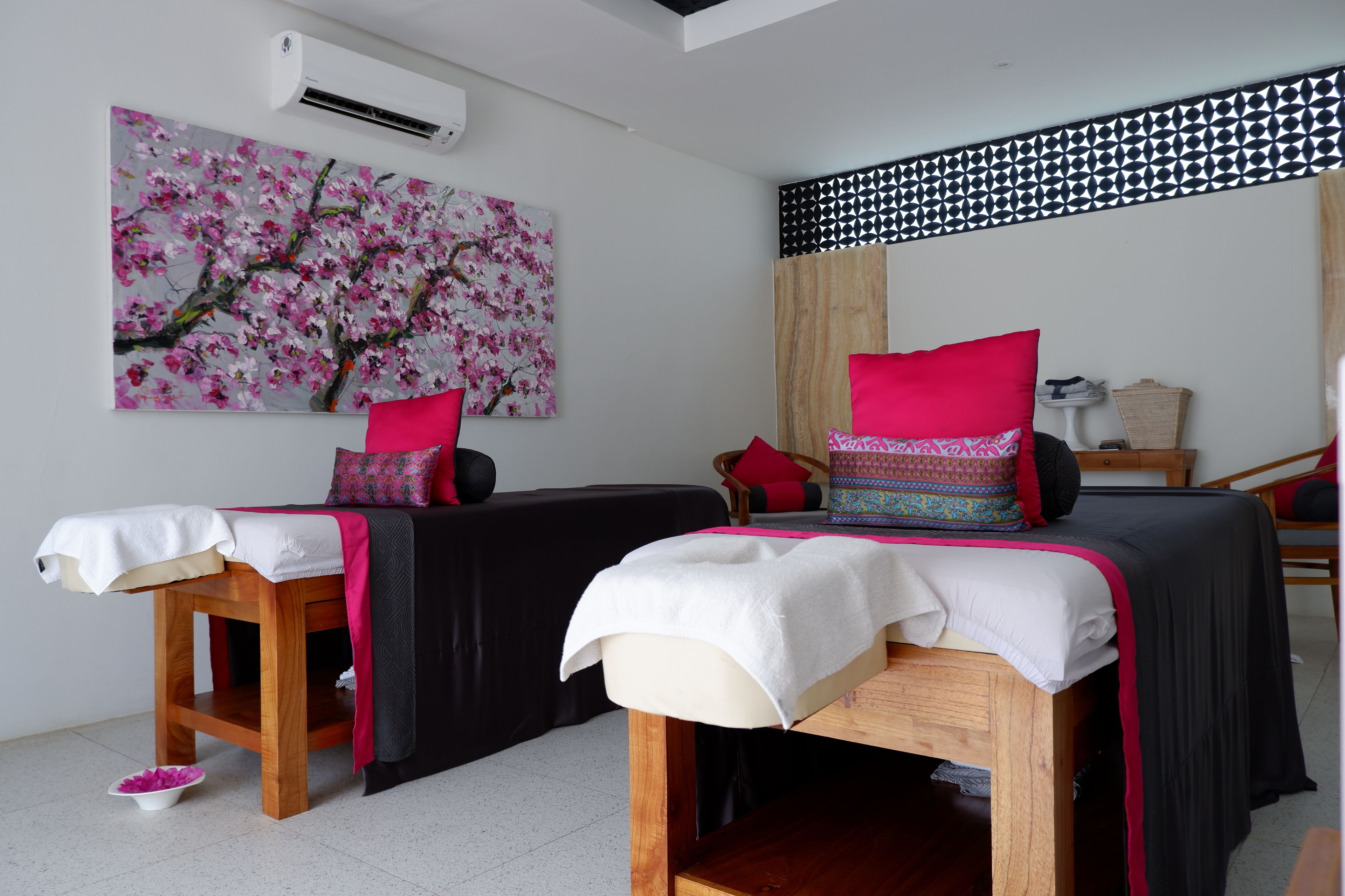 A room with two beds, a massage table, a wall-mounted air conditioner, and a large floral painting.