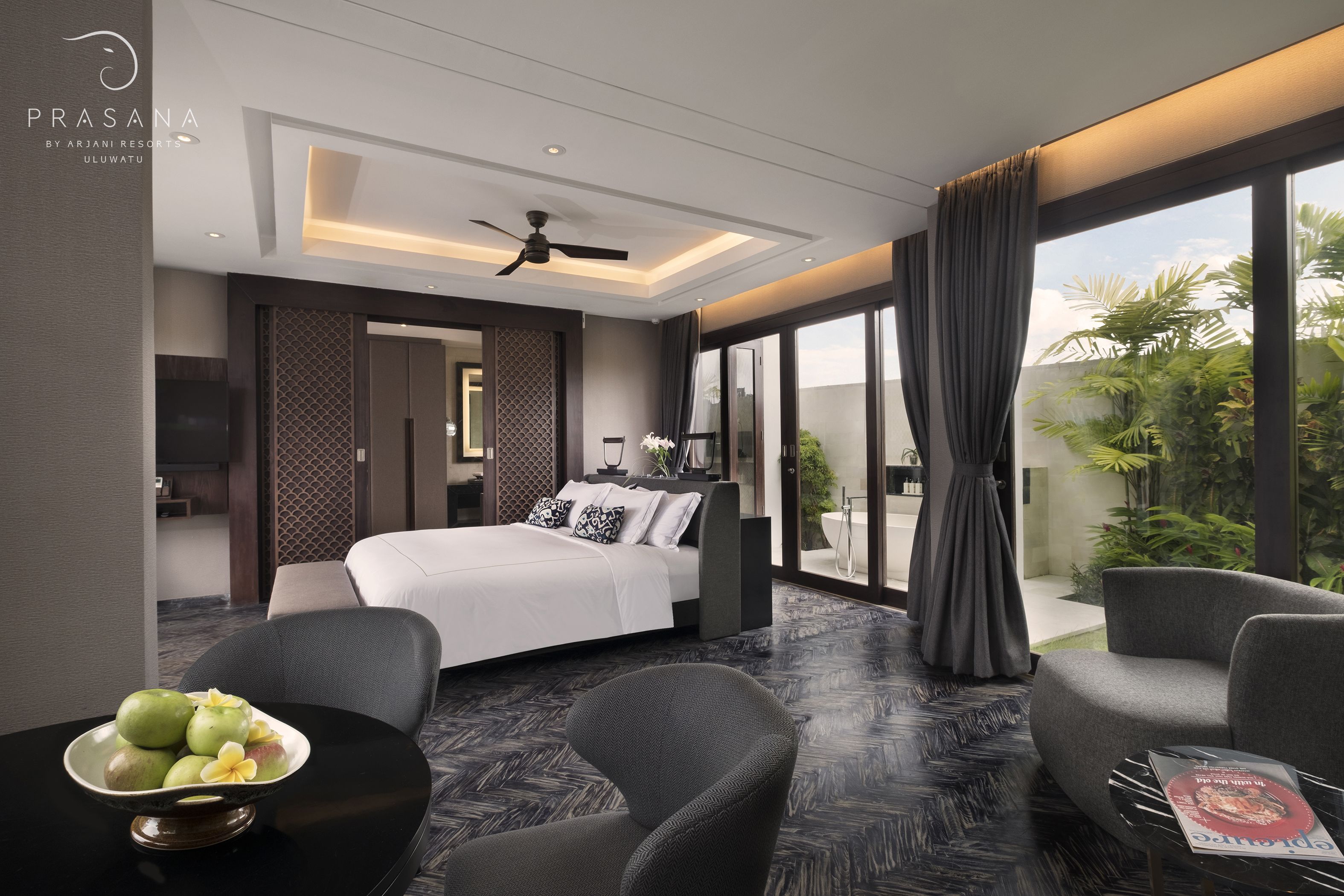 A modern hotel room with a bed, two chairs, a table with fruit, and a large window with a view of greenery.