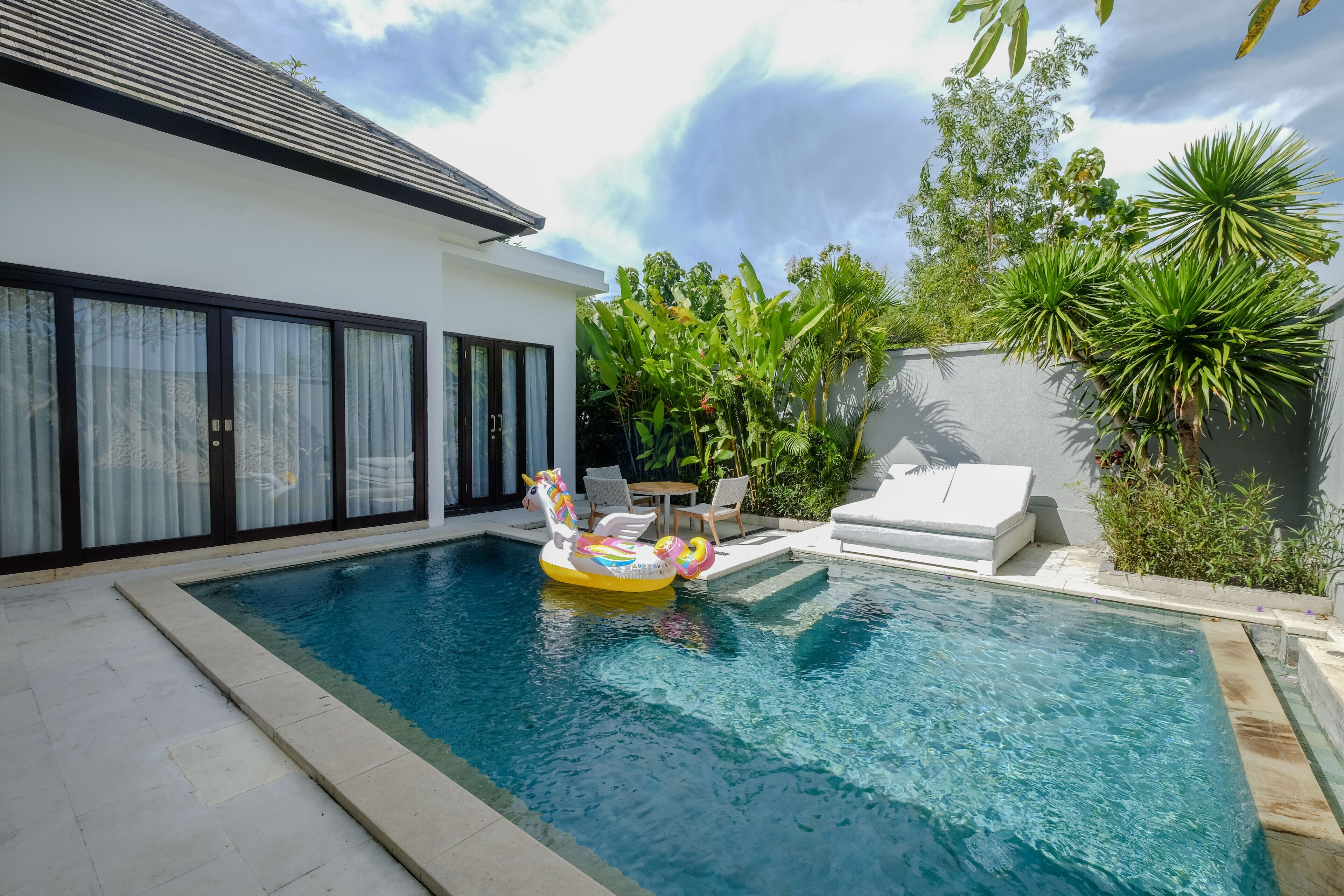 A swimming pool with a unicorn float, surrounded by a white house with large windows and a garden with tropical plants.