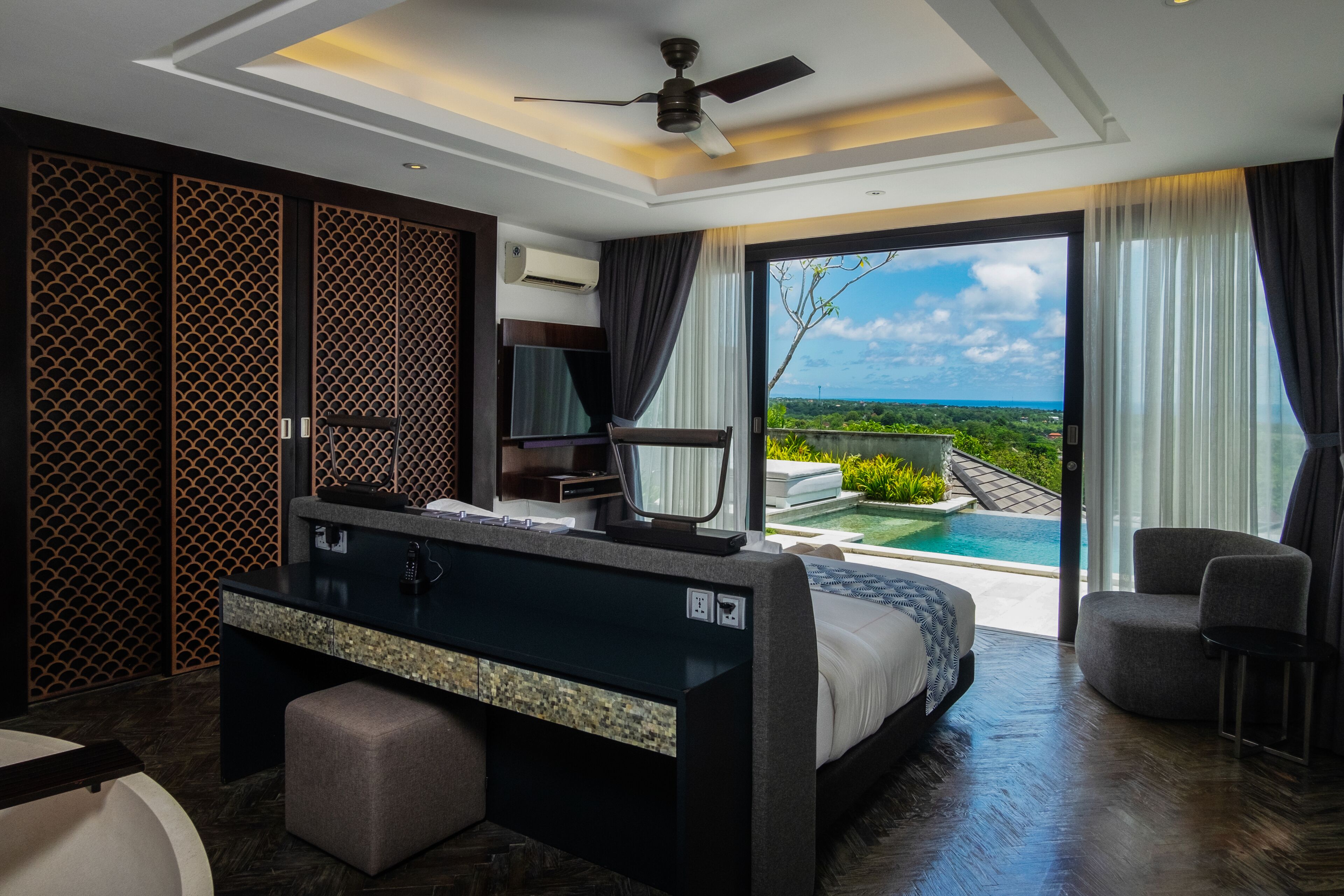 A modern bedroom with a bed, desk, and a view of a pool and landscape.