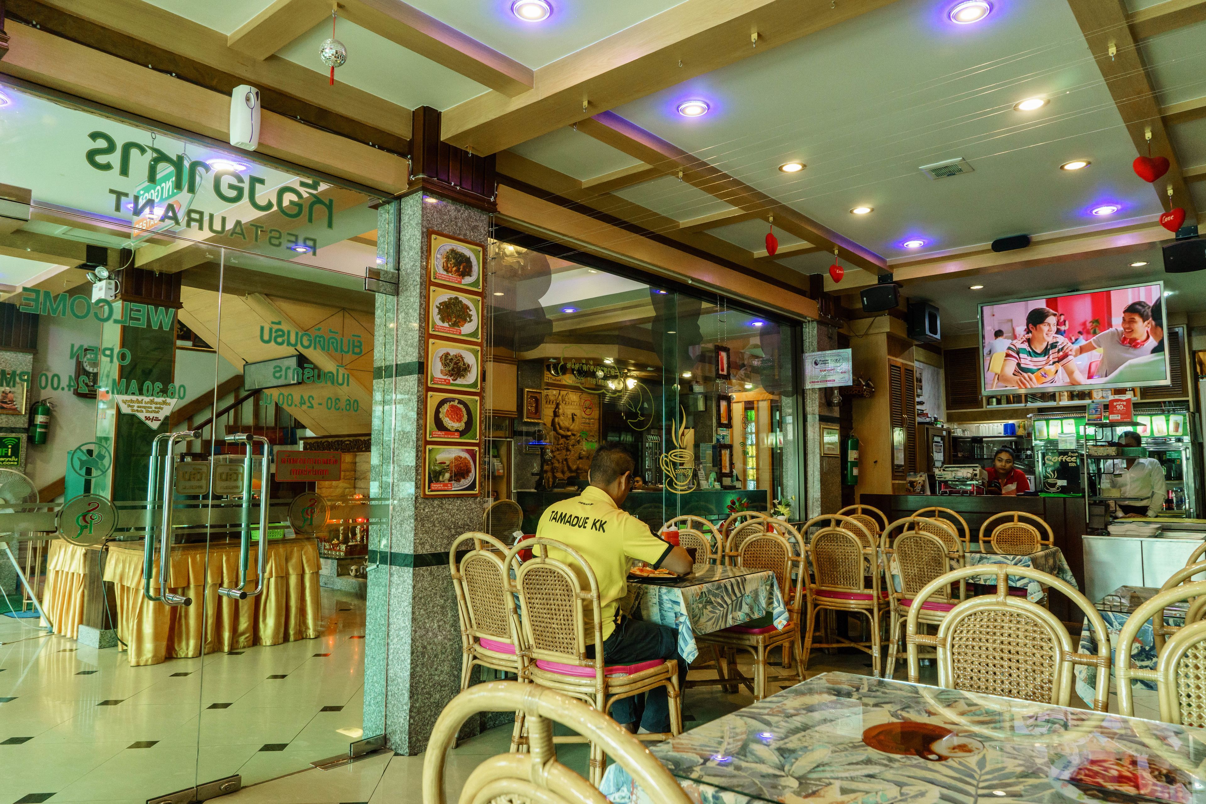 A restaurant with wicker chairs and tables, a counter with a person behind it, and a TV screen displaying a scene.