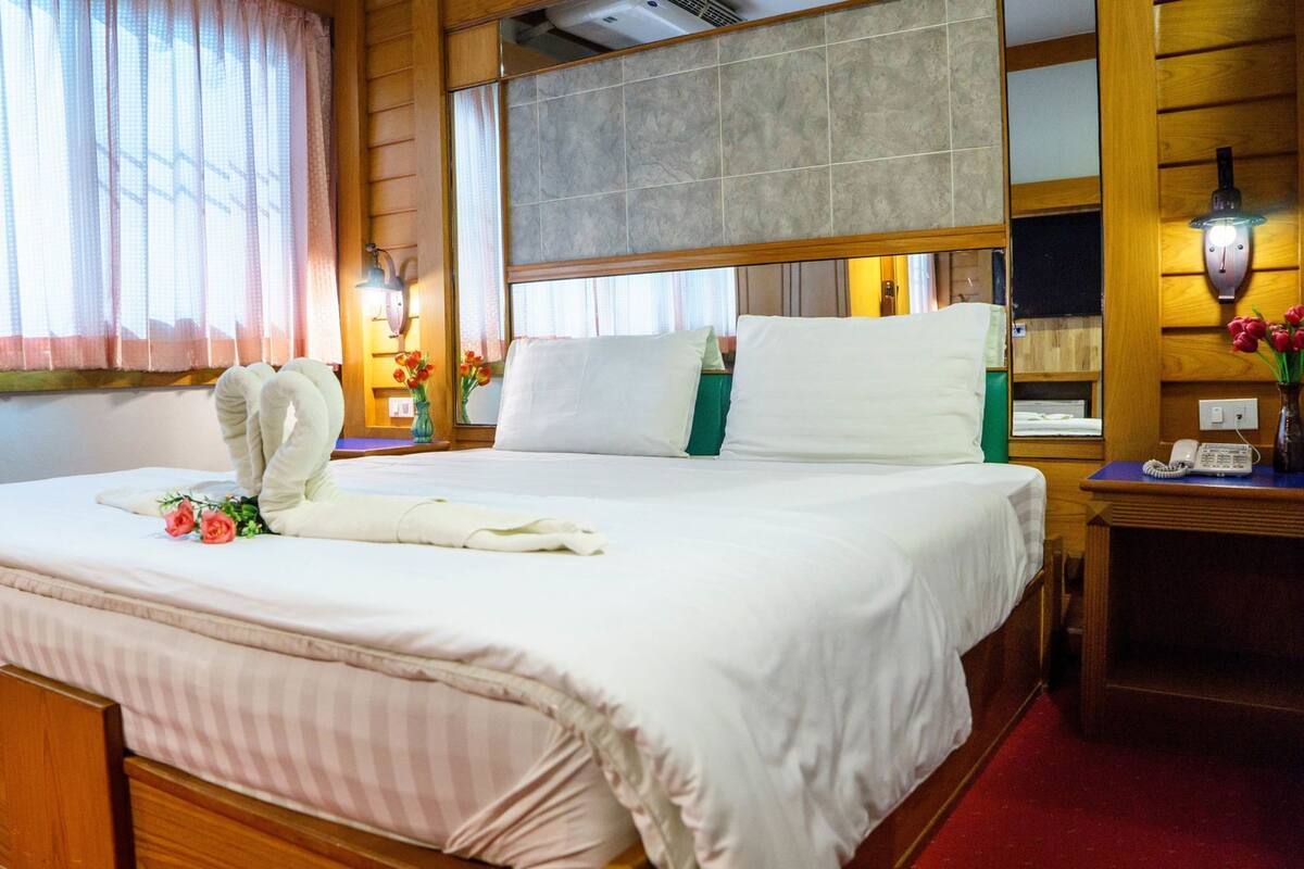 A neatly made bed with a towel and flowers, wooden paneling, a nightstand with a phone, and a window with curtains.