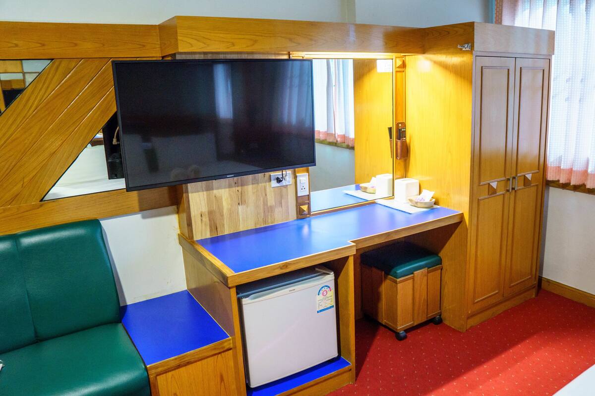 A compact hotel room with a wooden desk, a television, a small refrigerator, a green chair, and a wardrobe.
