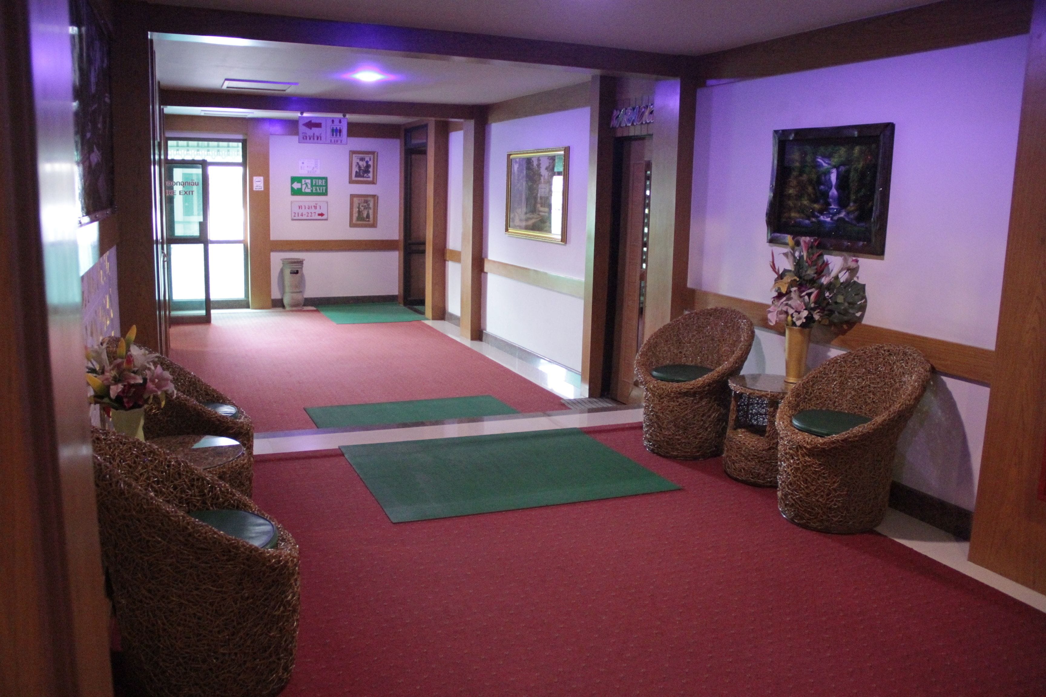 Un hall de hotel con sillones de mimbre, alfombra roja y felpudo verde. Hay cuadros enmarcados en las paredes y una planta en un jarrón.