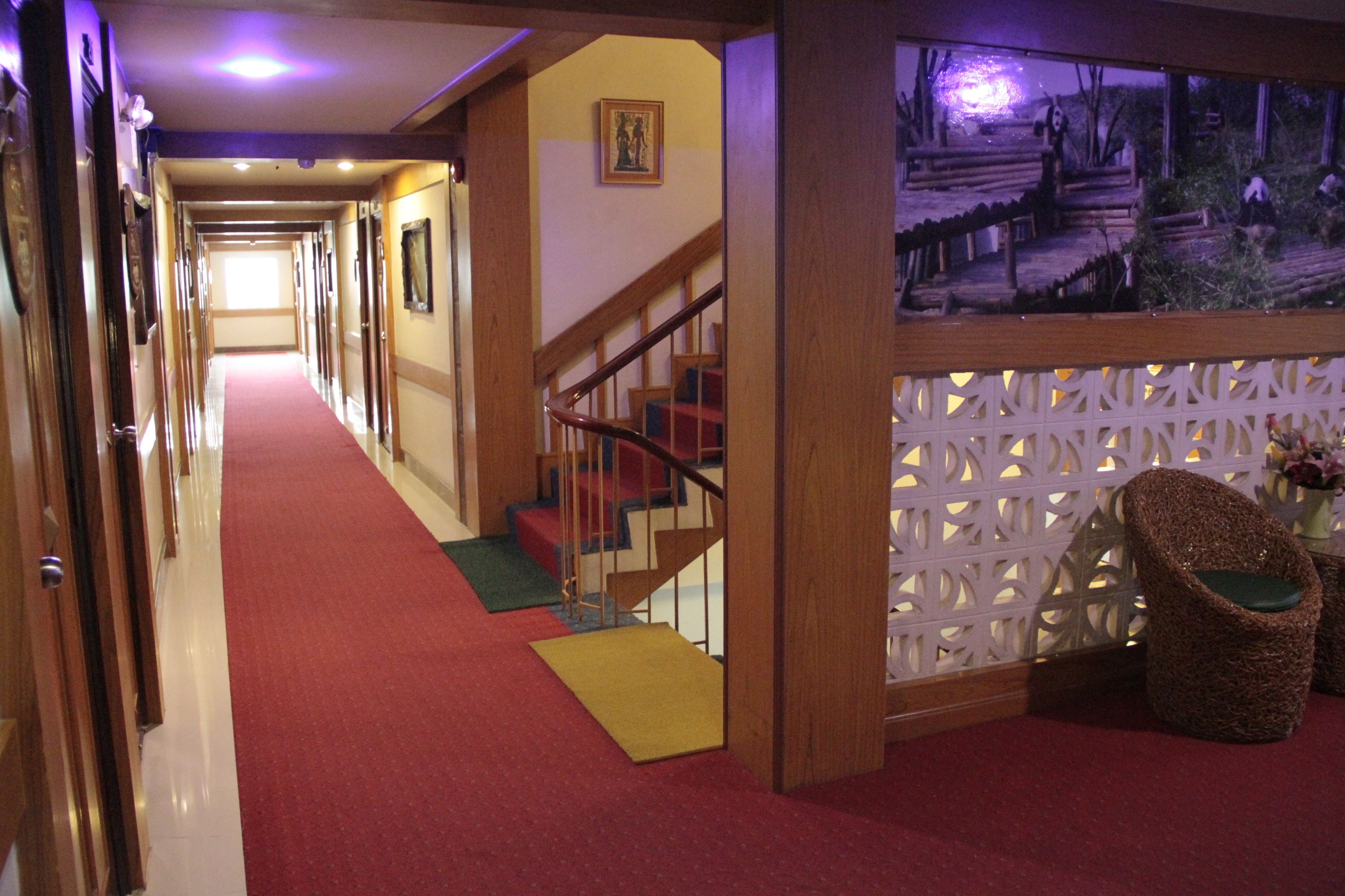 A hotel hallway with a red carpet, wooden walls, a staircase, and wicker chairs.