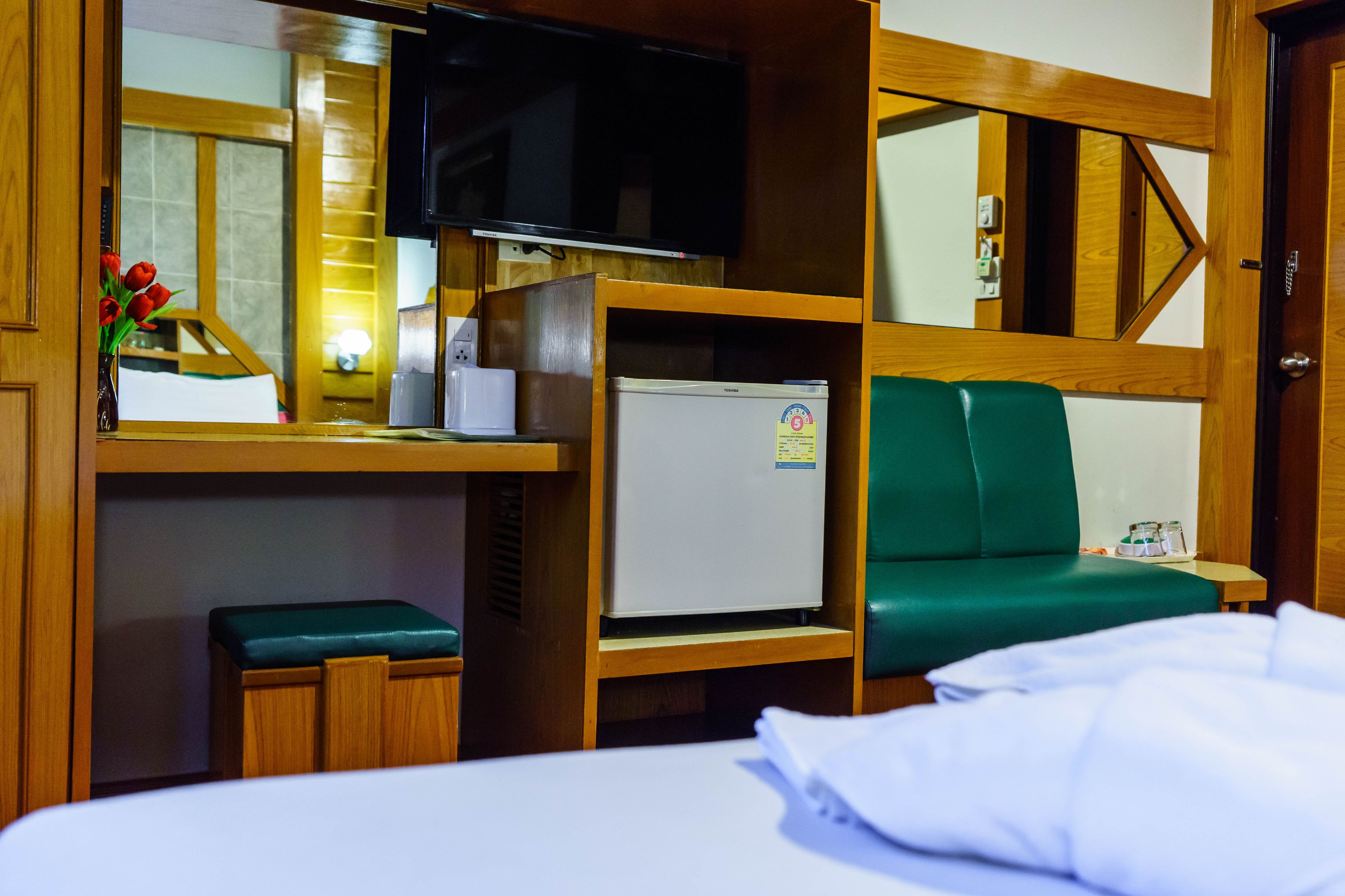 A compact hotel room with a bed, a small desk with a television, a refrigerator, and a green cushioned bench.