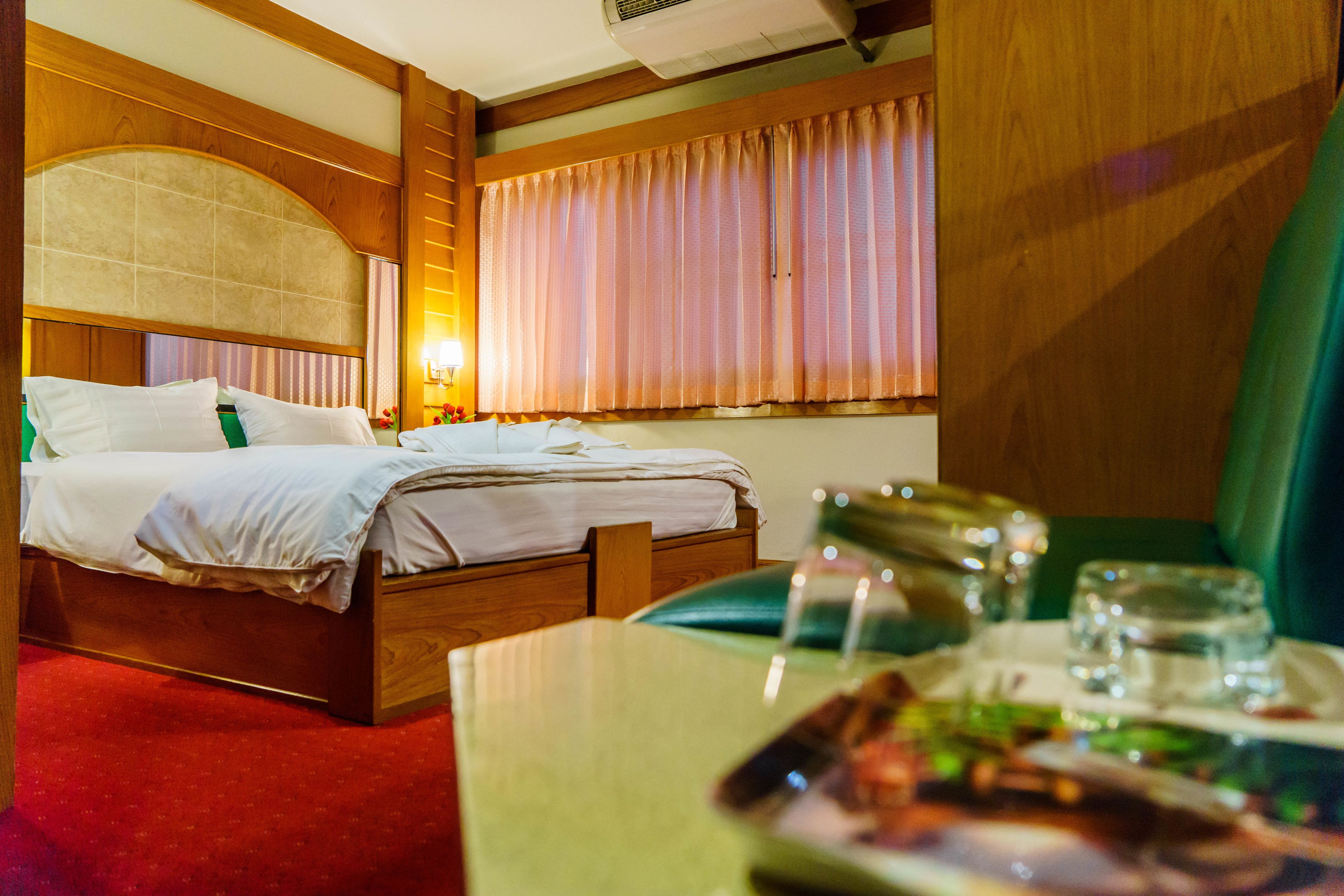 Habitación de hotel con una cama de madera, una mesita de noche con vasos y un plato de comida, y una ventana grande con cortinas.
