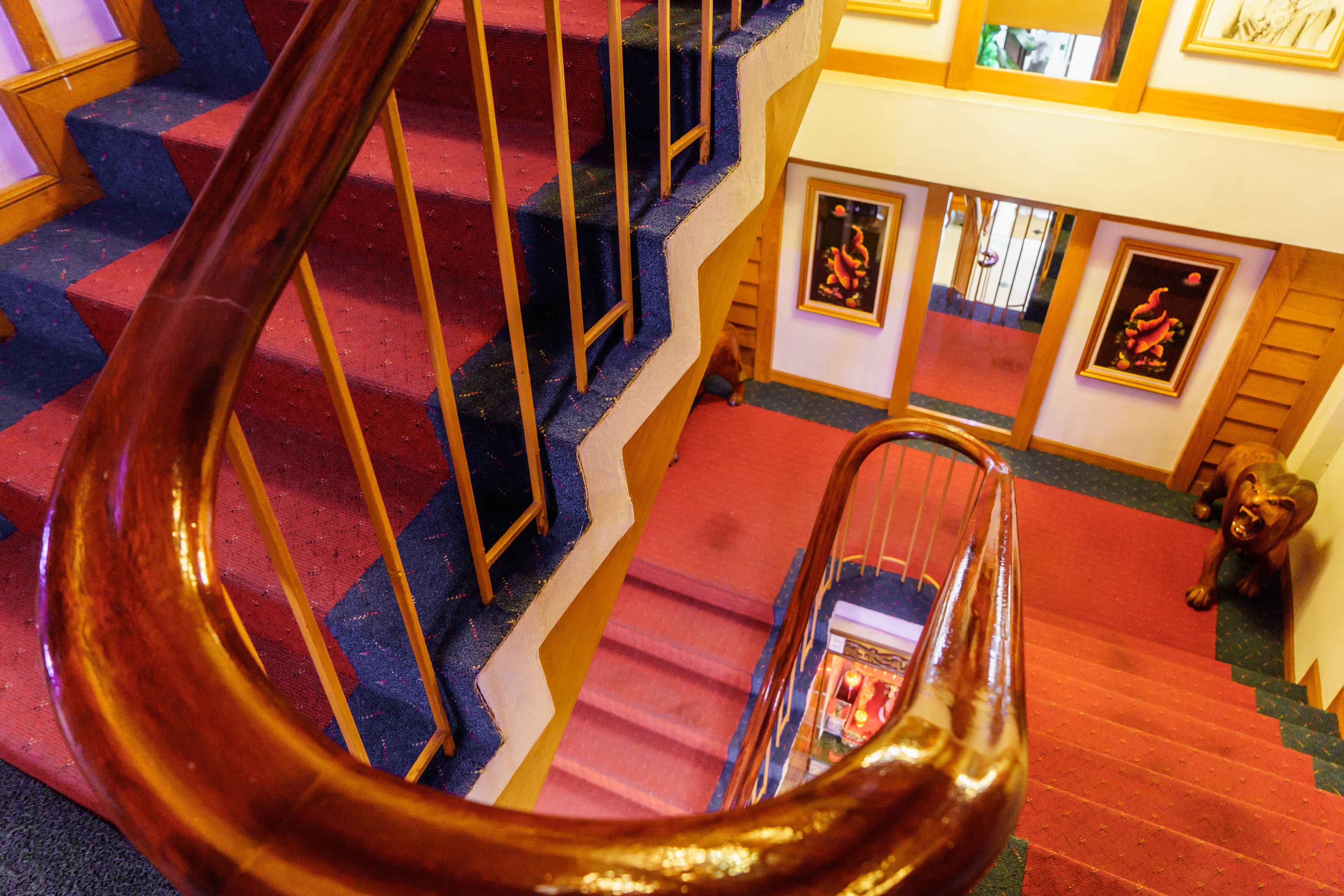 Una escalera de caracol con barandilla de madera, escalones alfombrados de color rojo y obras enmarcadas en las paredes.
