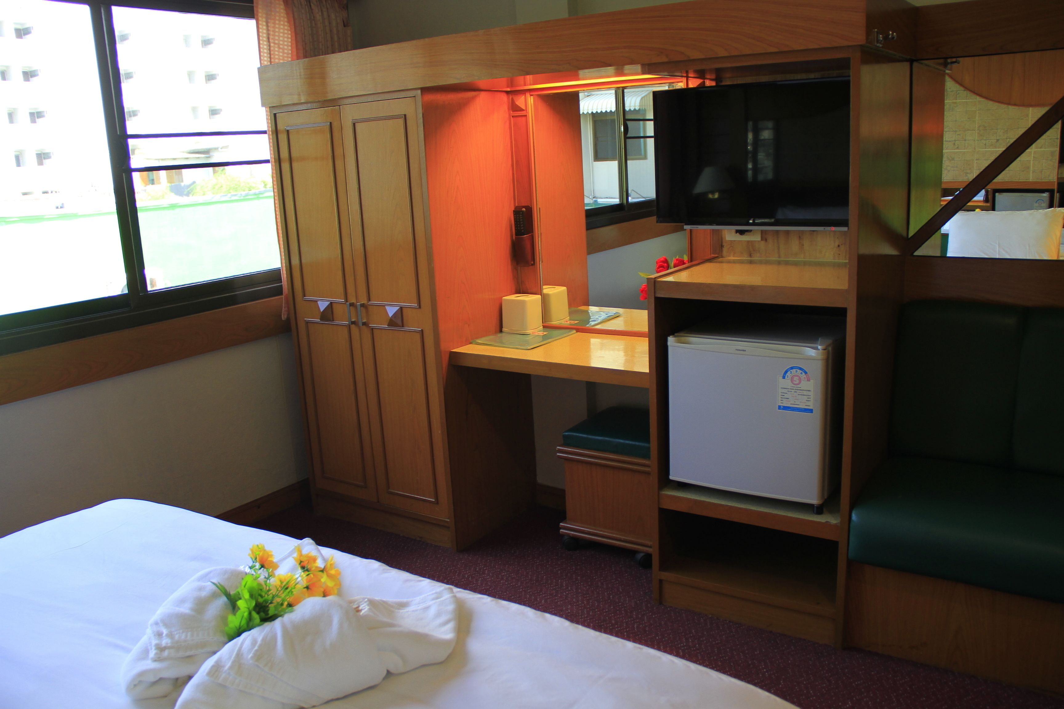 A hotel room with a bed, a desk with a television, a small refrigerator, and a window with a view of buildings.