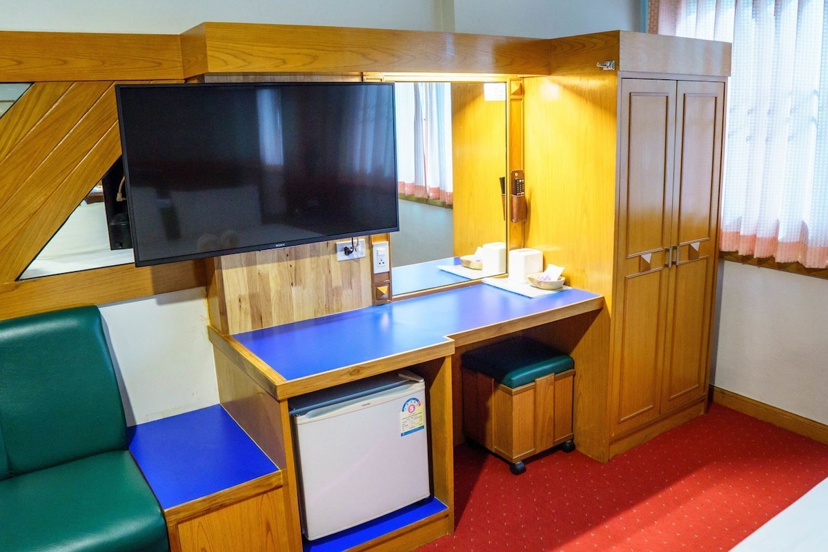 A compact hotel room with a desk, a television, a small refrigerator, a wardrobe, and a green sofa.