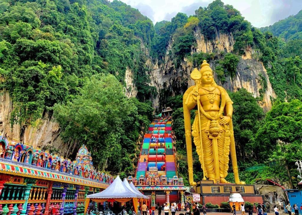 A large golden statue in front of a colorful temple complex with a steep, forested cliff in the background.