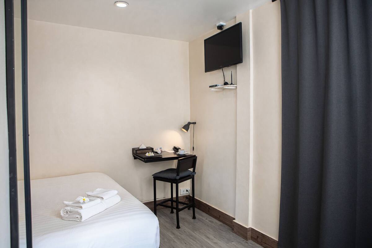 A hotel room with a bed, a desk with a chair, a flat-screen TV mounted on the wall, and a small shelf with a lamp.