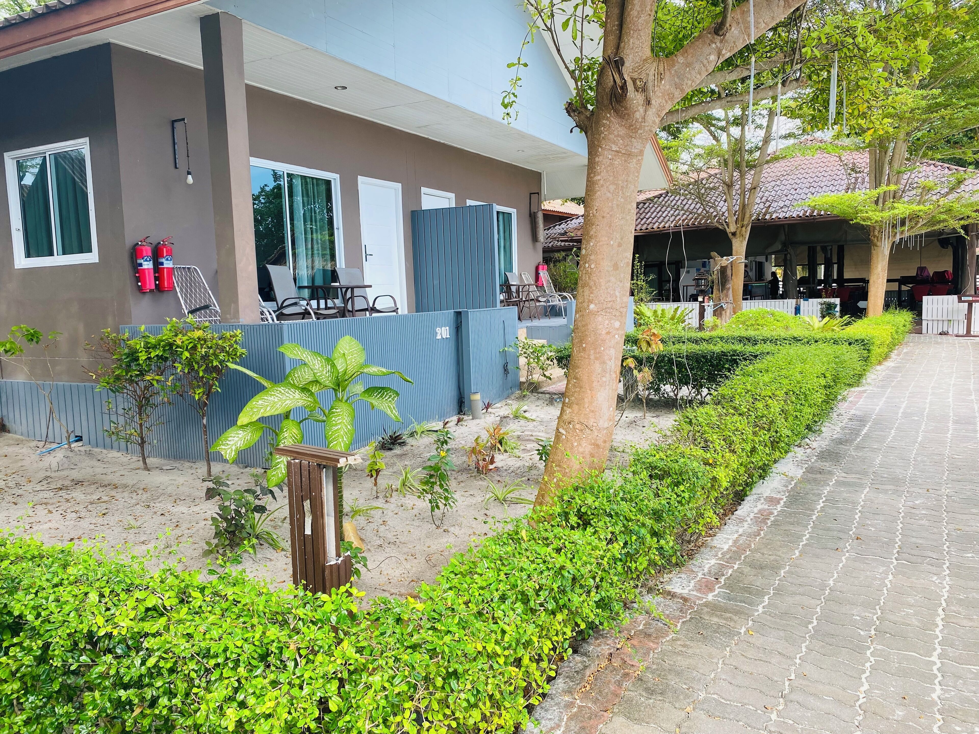 A building with a covered outdoor area, a paved walkway, and a fenced garden with various plants.