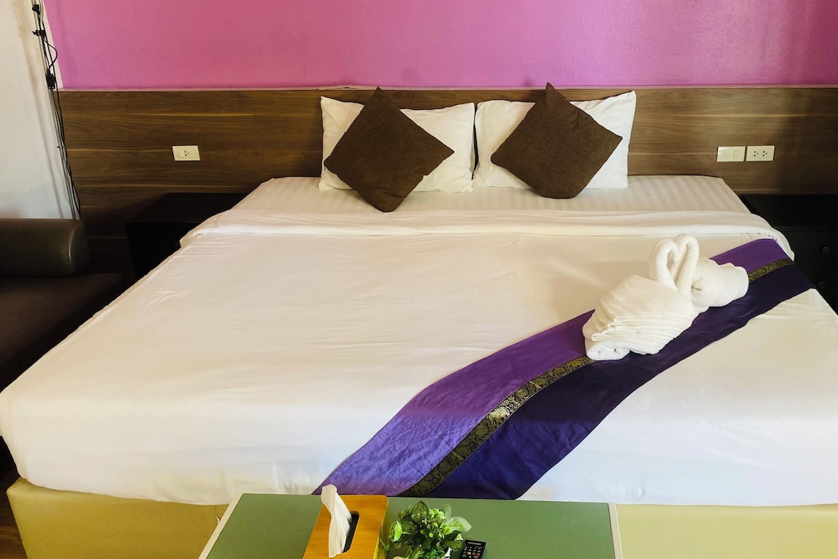 Een hotelkamer met een net opgemaakt bed, een decoratieve bedloper in paars en goud, en een witte handdoek gevouwen in de vorm van een zwaan op het bed.