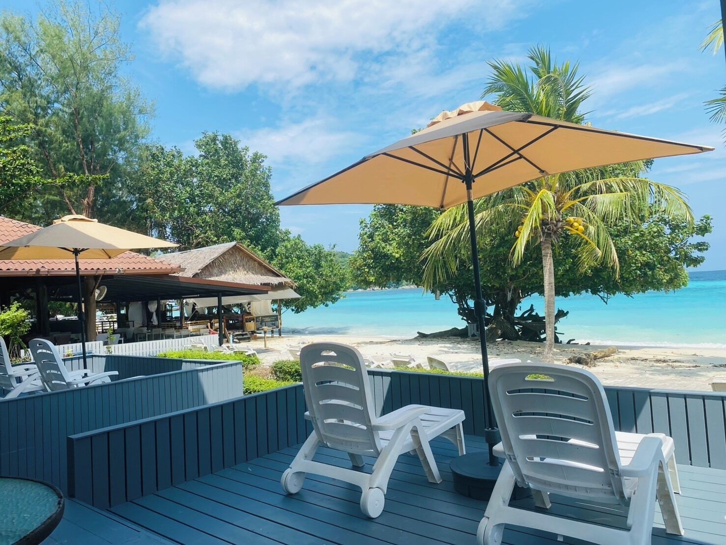 Une terrasse avec des chaises longues et une table, donnant sur une plage aux eaux bleu cristallin et bordée de palmiers.